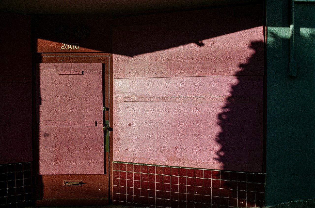Pink door with the number 2000 on top, surrounded by a pink wall, with shadows cast from an unseen object and some black tiles at the bottom.