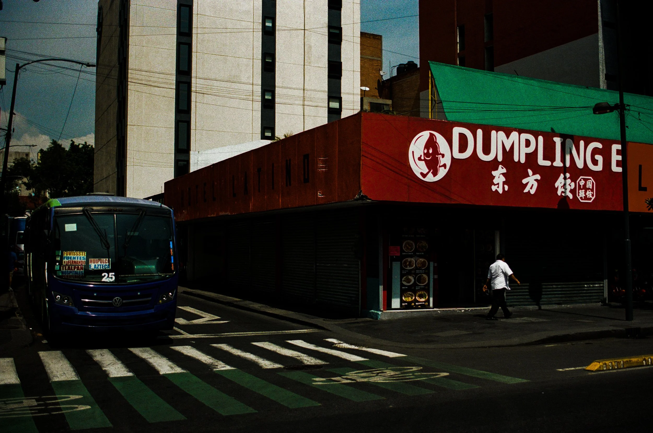 City street scene with a bus in the foreground, pedestrian crossing, and a red restaurant sign that says Dumpling. Tall buildings and power lines in the background.