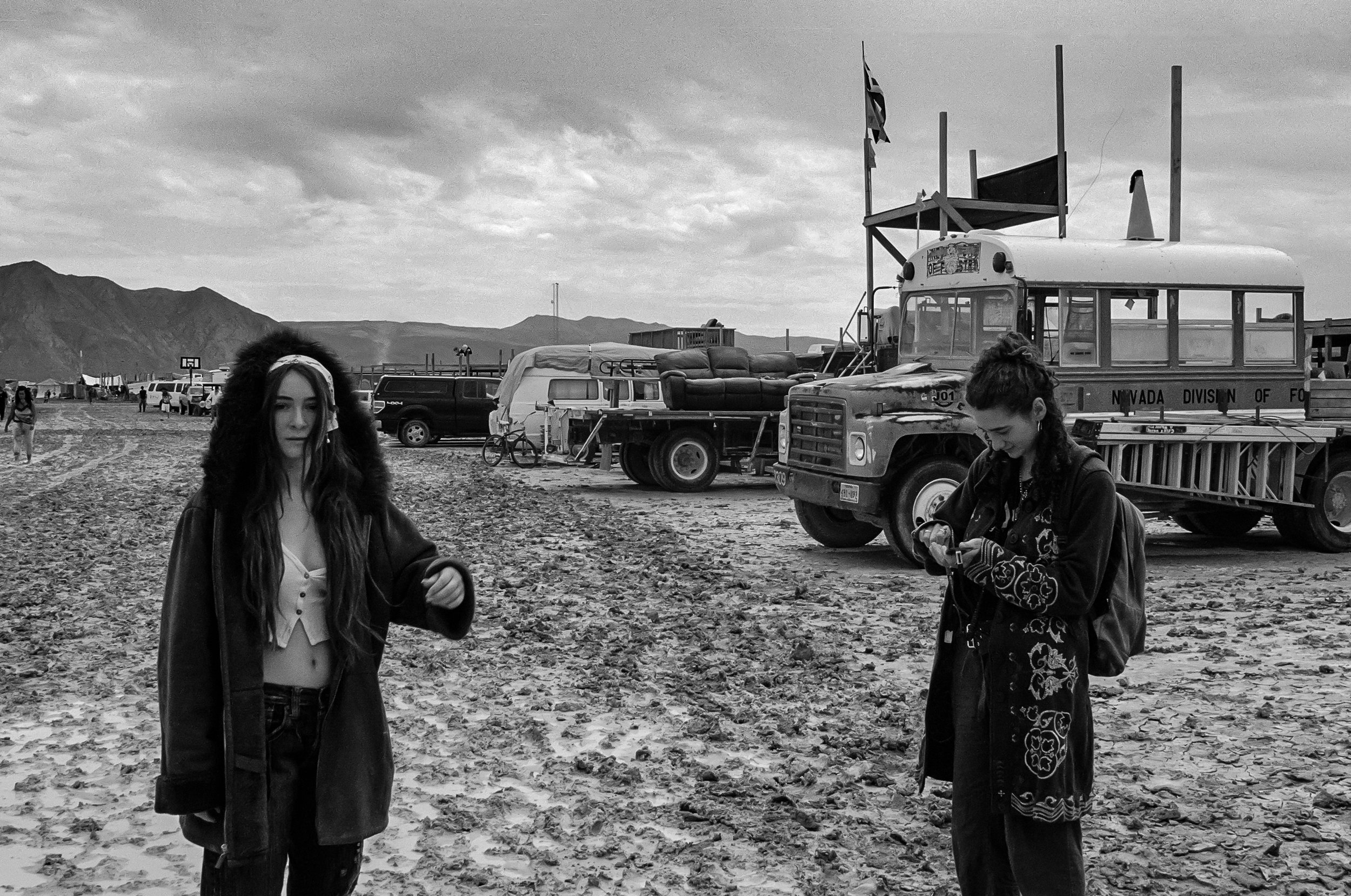 Black and white photo of a desert scene with two young women in the foreground, one with long hair and the other with her hair tied back, standing on rocky ground. In the background, there are vintage trucks, chairs, and flags, with mountains and a c