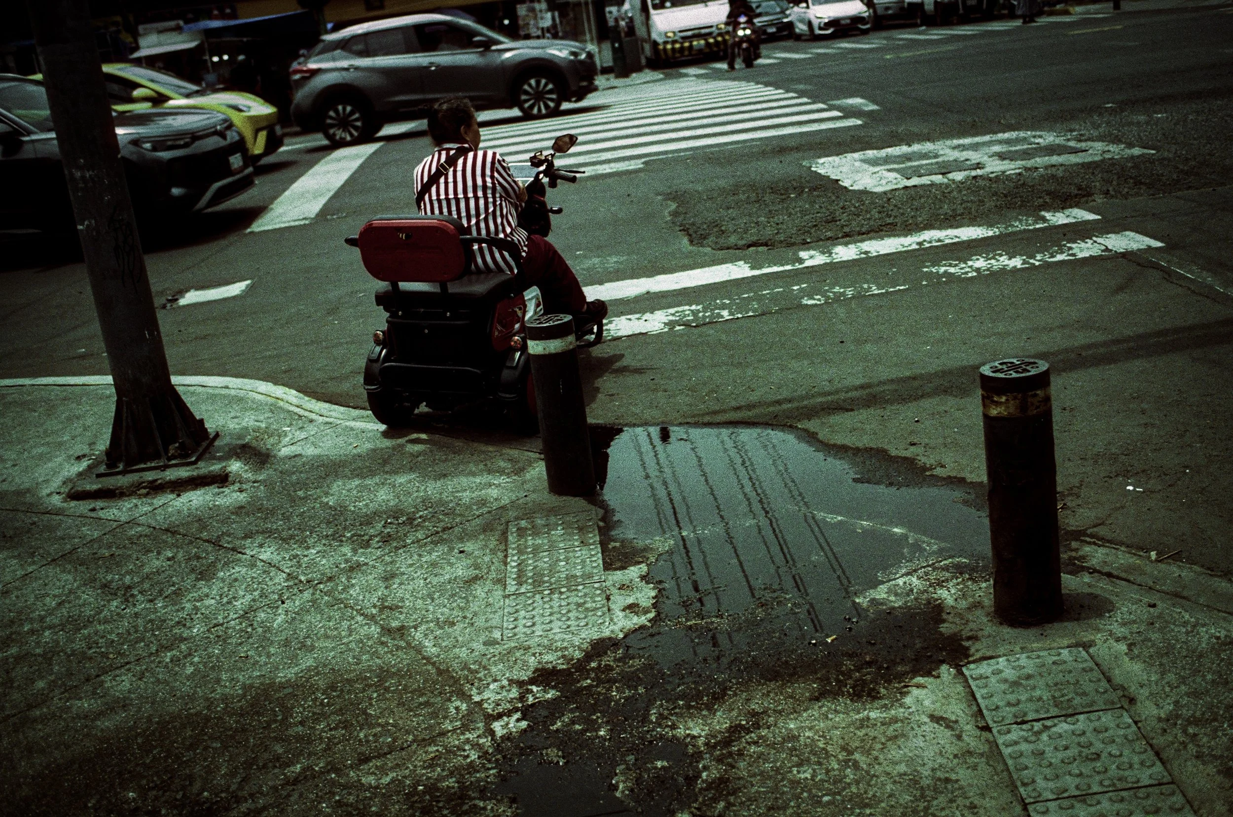 A person on a mobility scooter waiting to cross a city street at a crosswalk, with parked cars and a bus in the background, and a wet street surface.