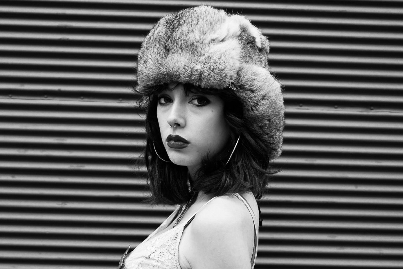 Black and white portrait of a woman with dark wavy hair wearing a fluffy fur hat, hoop earrings, and a white lace top, standing in front of a striped metal background.