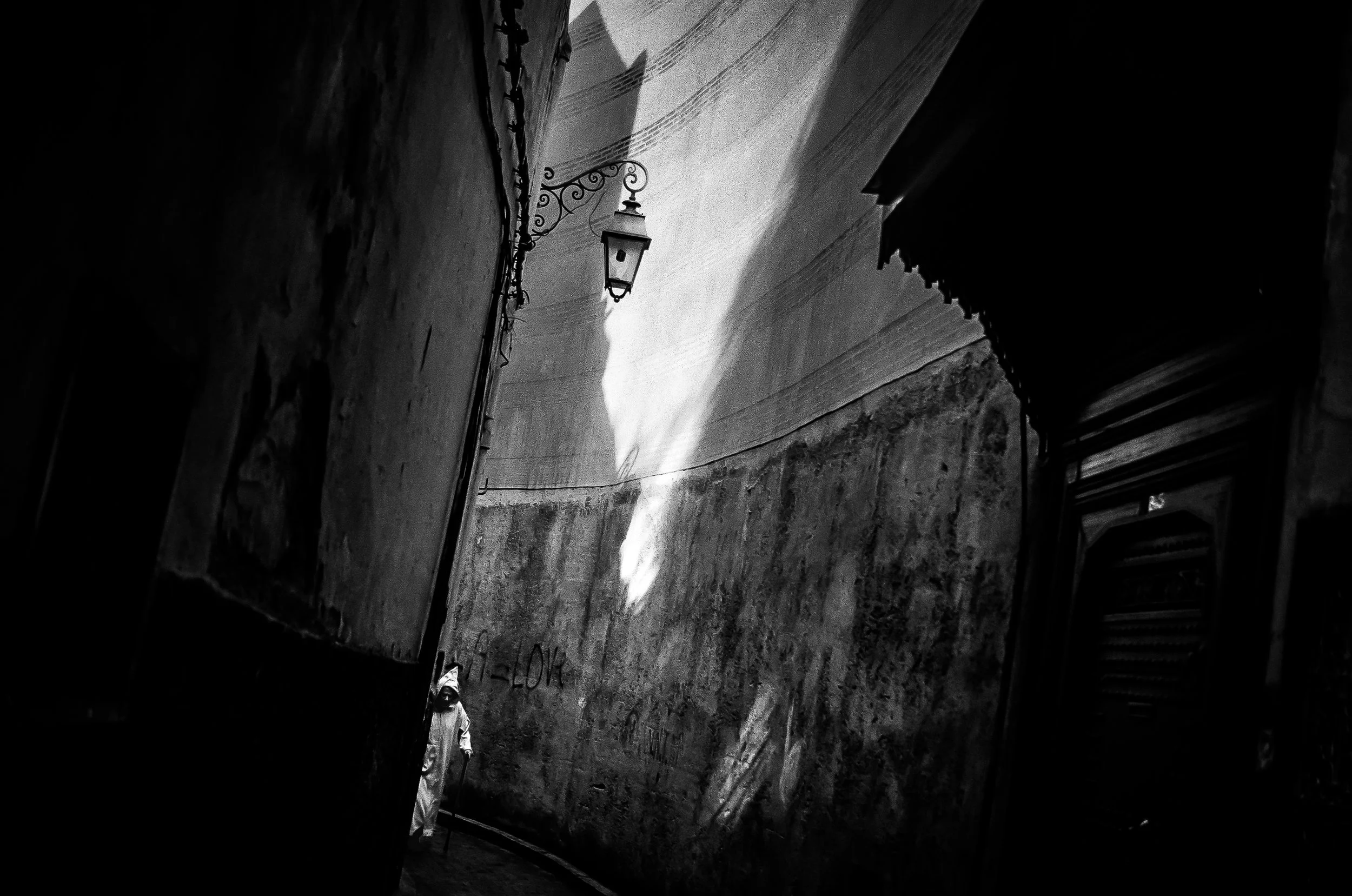 A narrow alley with textured stone walls, an ornate vintage street lamp attached to the left wall, and graffiti on the bottom of the wall. The photo is in black and white.