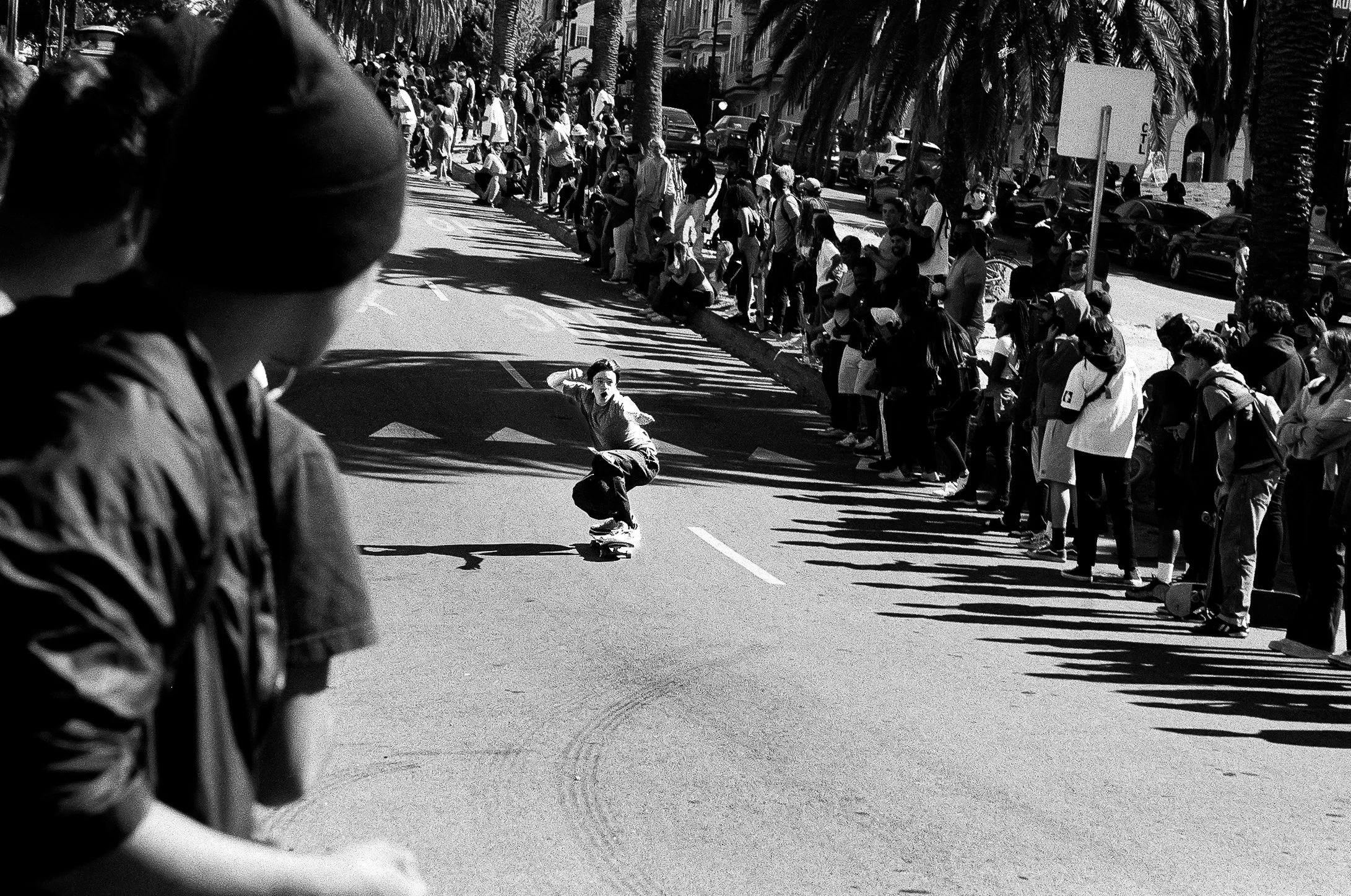 People standing in line along a sidewalk, with a skateboarder in the foreground, casting a long shadow, in a city street lined with palm trees.