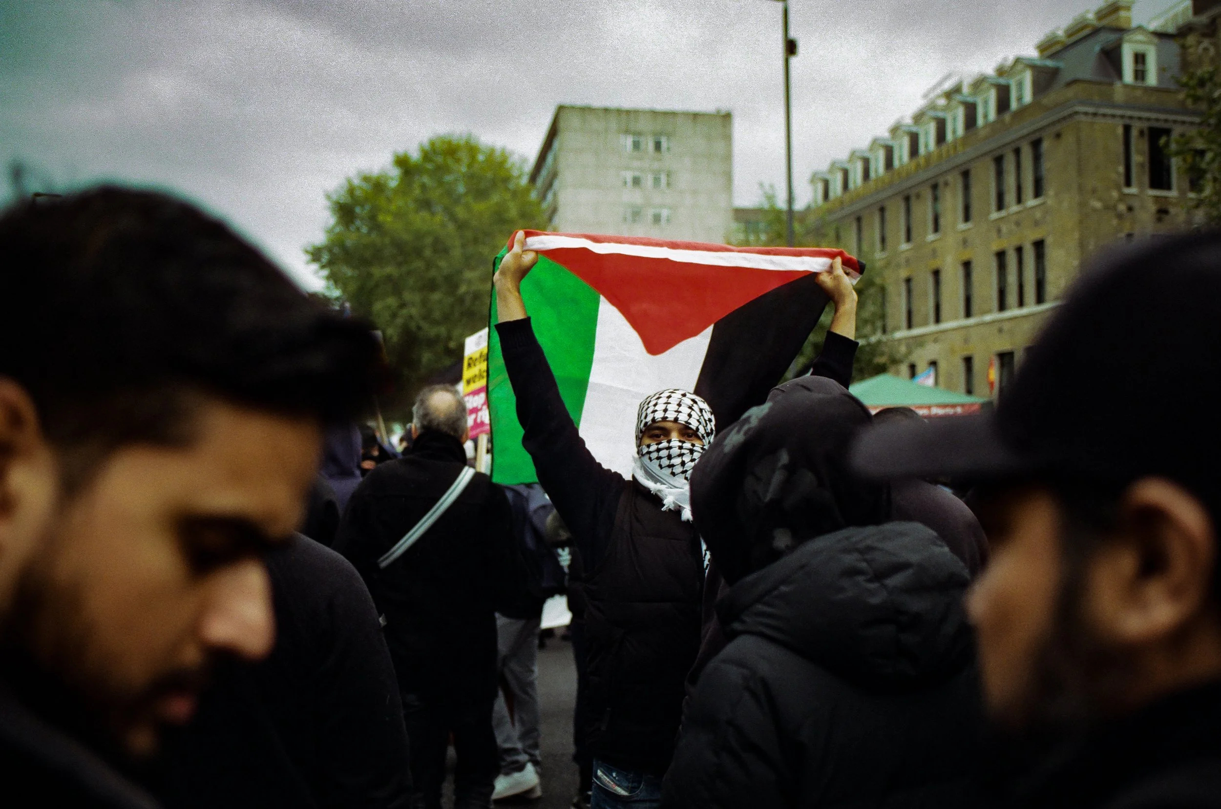 A crowd of people gathered outdoors, with one person holding a flag that appears to be the Palestinian flag. The person is wearing a black jacket and a black-and-white patterned headscarf. The scene takes place in an urban area with buildings and tre