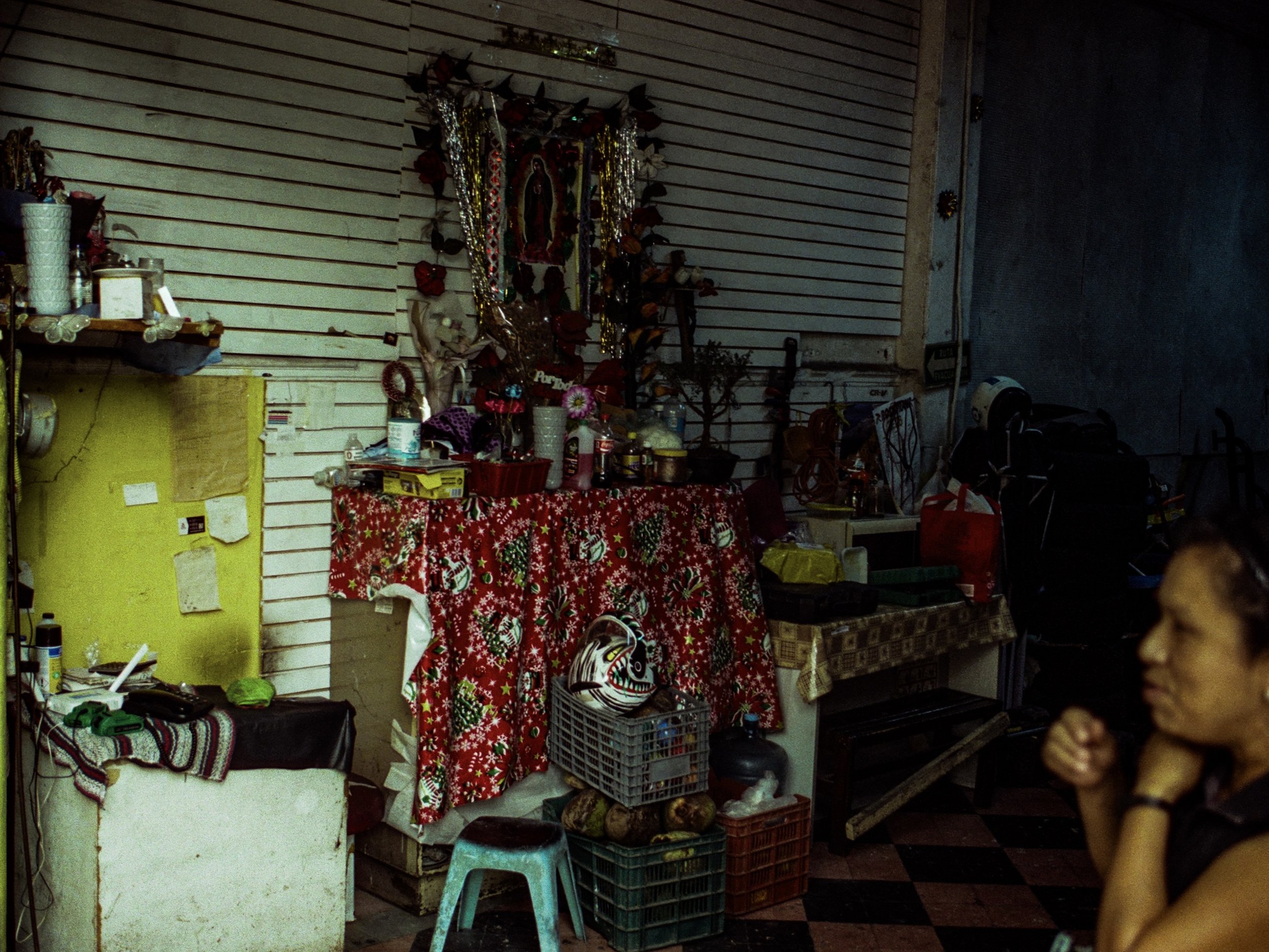 A cluttered room with food, drinks, and various decorations on the table covered with a red and white Christmas-themed tablecloth, with other items such as plastic crates, a chair, and a small sofa visible, and a religious image and garlands hanging 