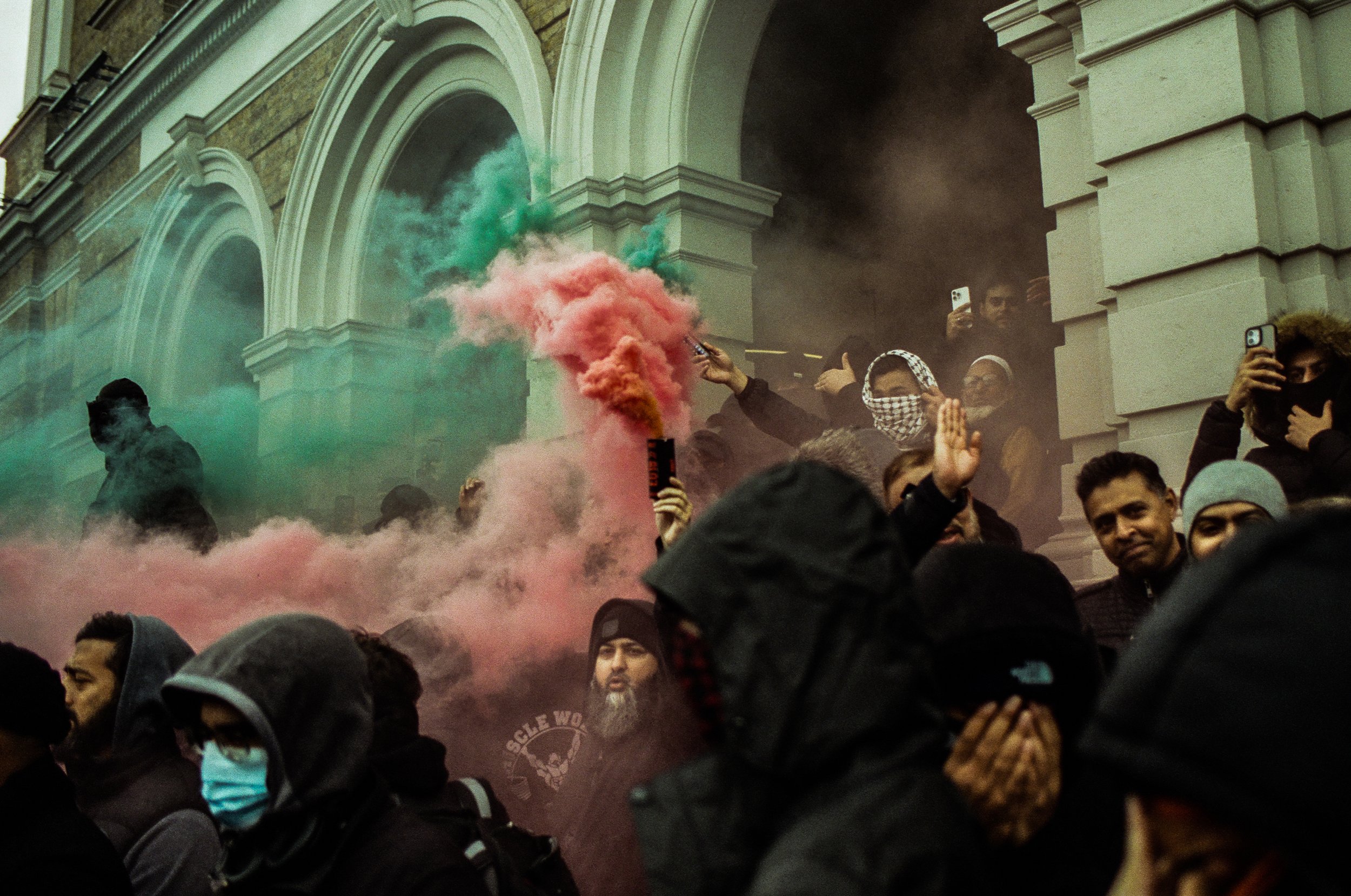 A crowd of people gathered outside a building, with some holding phones and a person holding a smoke flare emitting pink and green smoke, with people wearing face masks and head coverings.