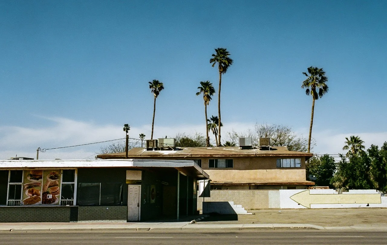 A single-story building with a brown roof and a larger, two-story beige building behind it, with tall palm trees in the background under a blue sky.