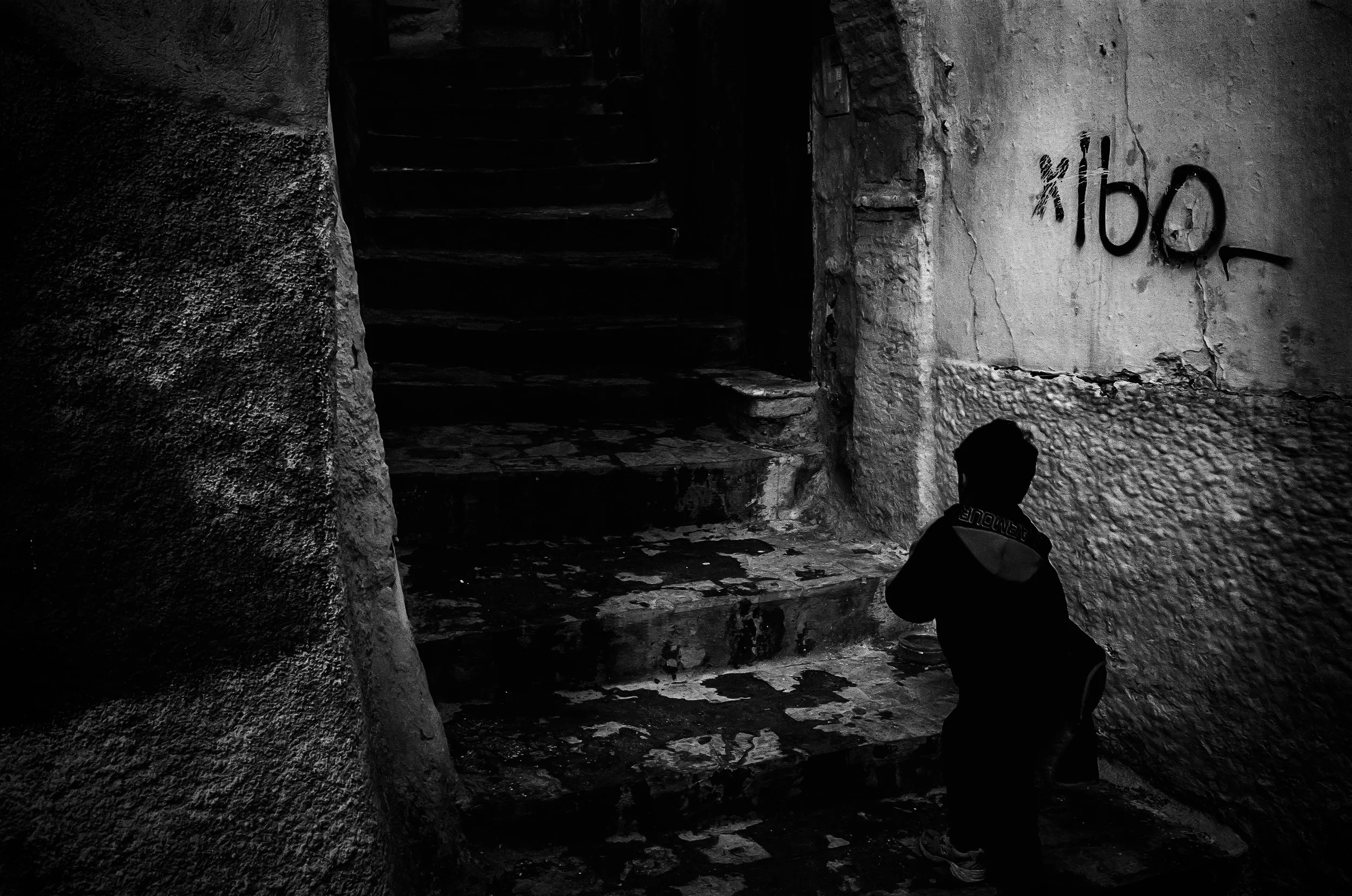 A person standing inside a dark, narrow stone hallway or stairwell with textured walls and steps leading upward. The person is wearing a jacket with text on the back and is partially visible at the lower right of the image. There is graffiti on the w
