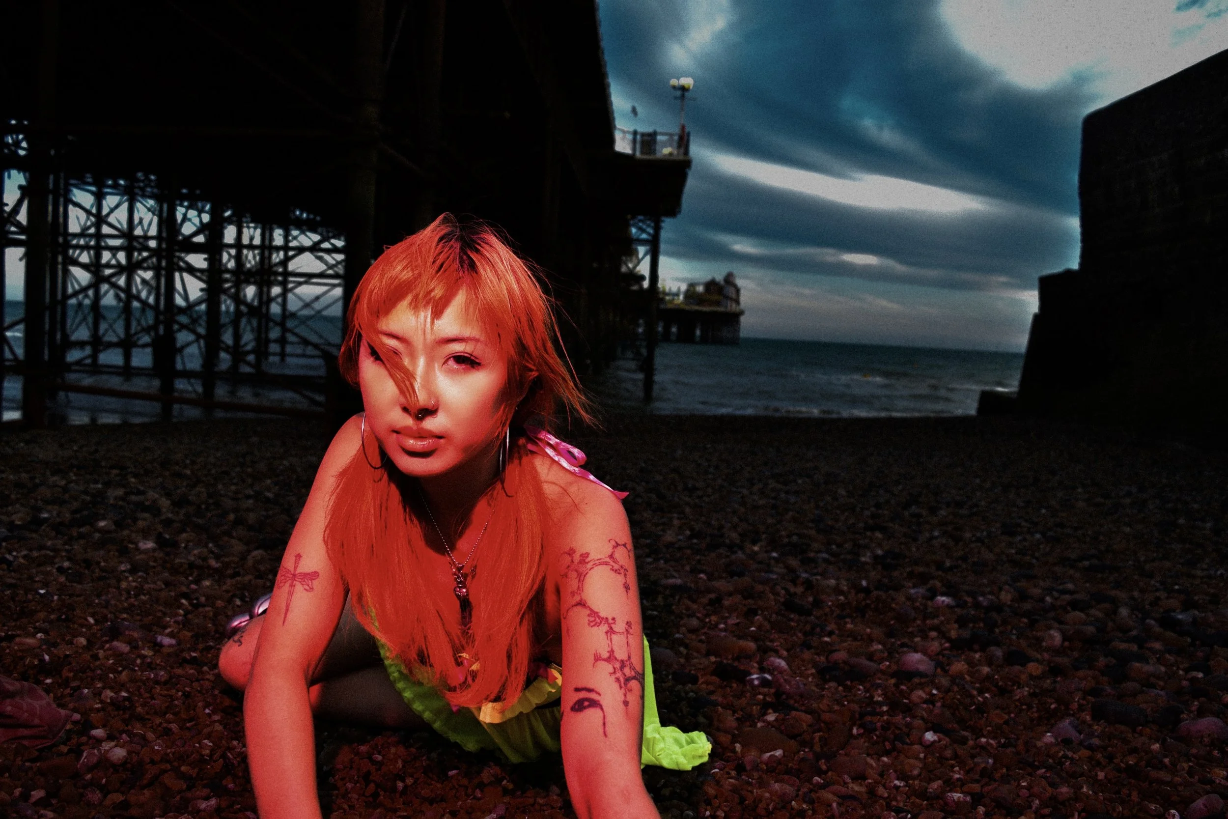 A woman with bright orange hair, tattoos, and piercings crouches on a dark rocky beach near a pier with an industrial structure, under a cloudy sky at dusk.