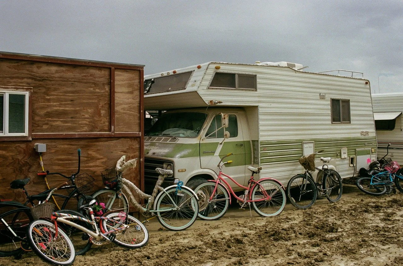 An old, beige and green motorhome parked next to a wooden building. Several bicycles are lined up in front of the motorhome, some with baskets and flowers, on a muddy ground under an overcast sky.