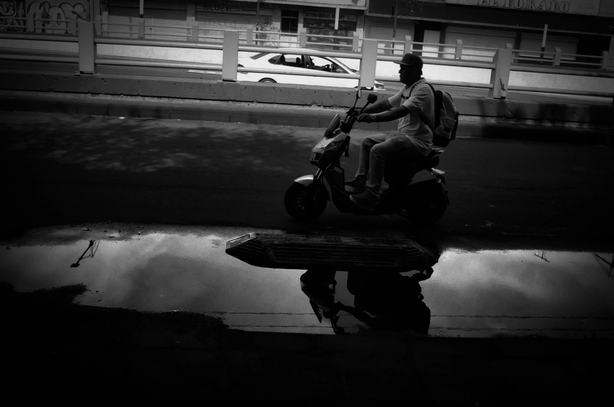 A man riding a scooter on the street with a backpack, wearing a helmet, and reflected in a puddle on the ground, with vehicles and buildings in the background.