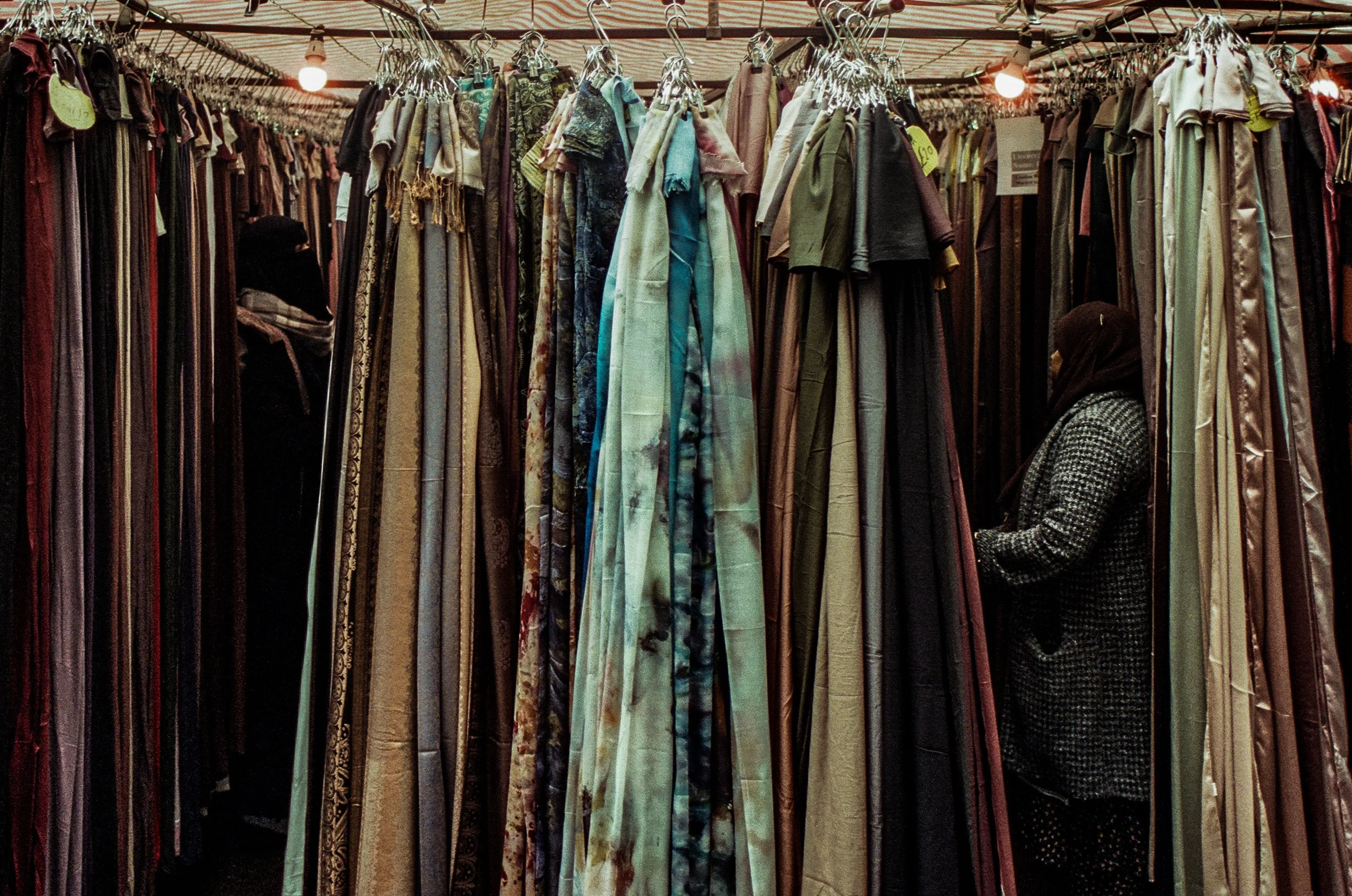 A market stall with hanging clothing, including pants, dresses, and shirts in various colors and patterns. Two women wearing headscarves are browsing the clothes.