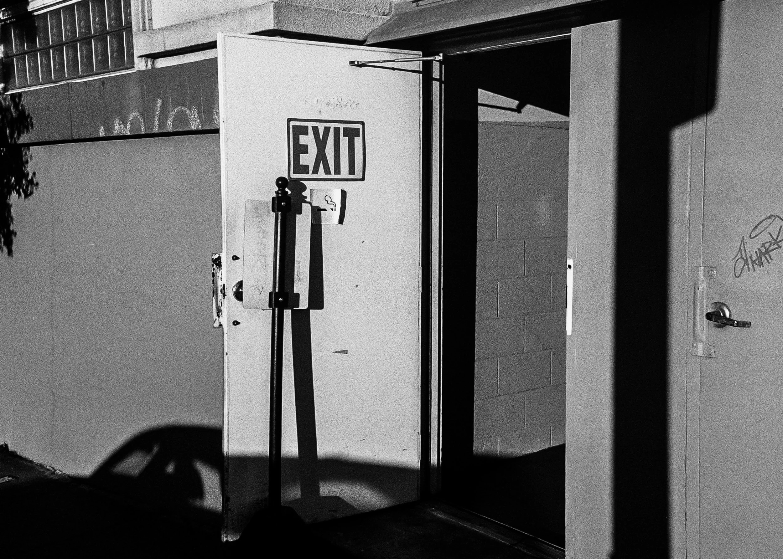 A black and white photo of an exit door with a sign. The door is open, revealing a dark interior. There is a small sign with a cigarette symbol and a line through it, indicating no smoking.