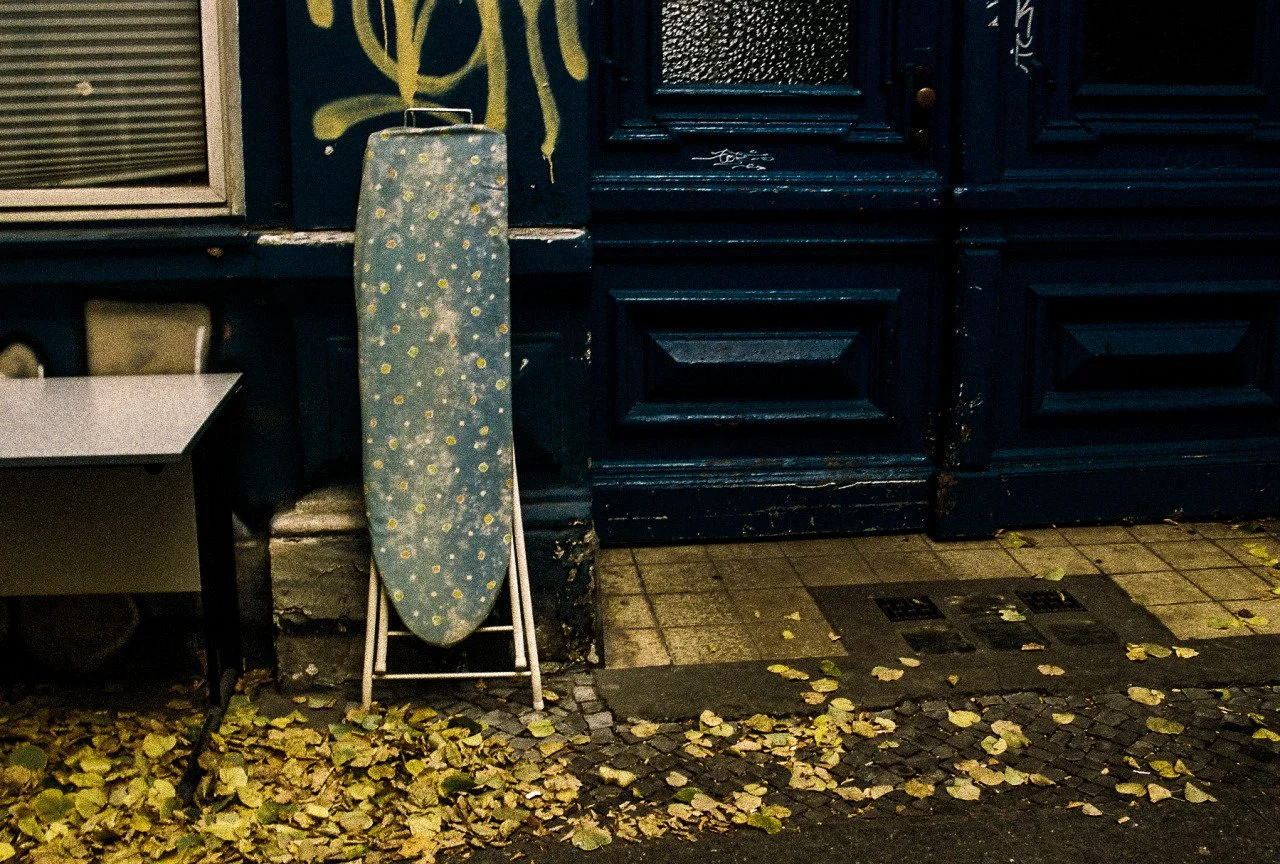 An ironing board with a blue cover decorated with yellow flowers leaning against a dark blue door. The ground is covered with fallen yellow leaves.