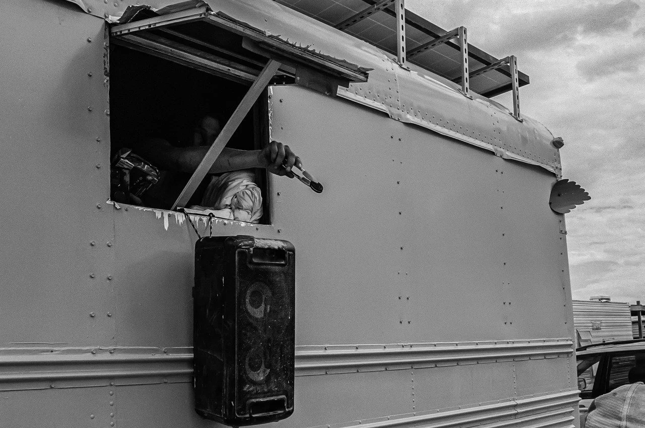 A person inside a food truck speaking into a microphone, with a large speaker mounted on the exterior wall. The food truck has a fish-shaped decoration on the corner. The image is in black and white.