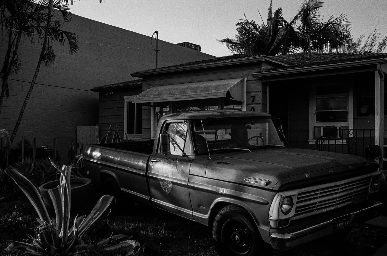 A vintage pickup truck parked in front of a small house with a porch, surrounded by plants and palm trees, in black and white.