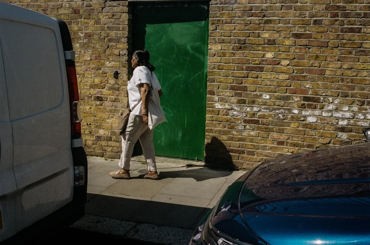 A woman with dark hair wearing a white shirt and light-colored pants walking on the sidewalk in front of a brick wall with a green door, next to a parked white van and a dark-colored car on a sunny day.