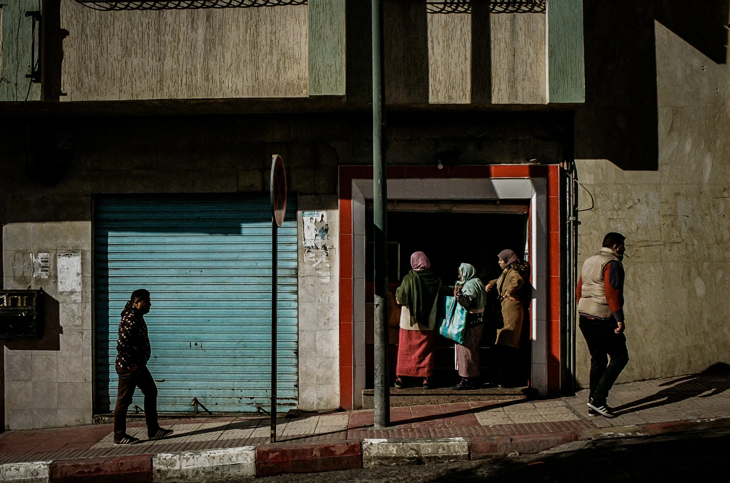 People walking outside a building with a red and gray entrance, a blue roller shutter door, and posters on the wall. Shadows cast by a streetlight or sign.