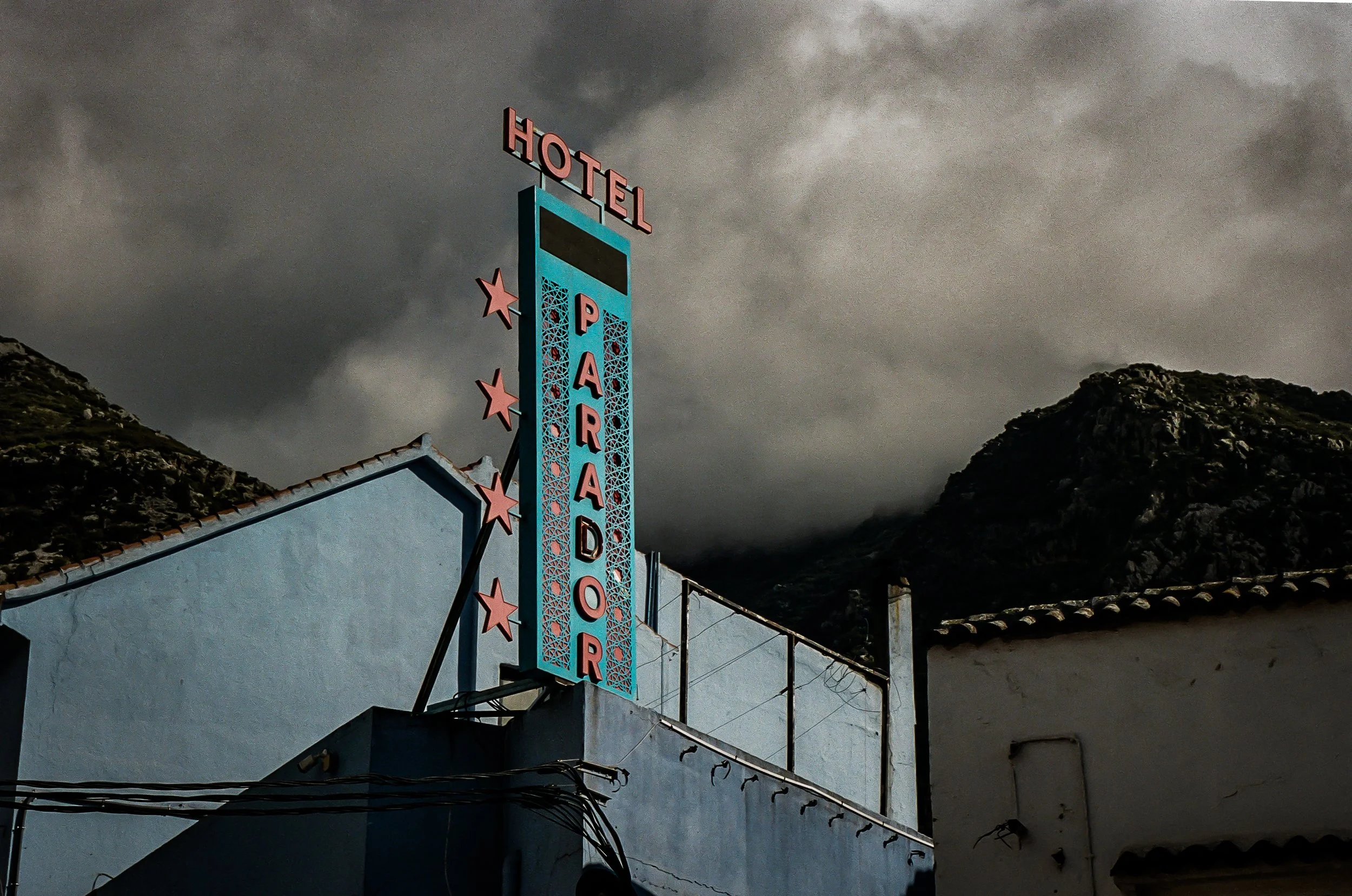 A vintage sign for a hotel with a blue and pink color scheme, featuring a tall vertical sign with pink stars and letters, set against a dark, cloudy sky and mountainous landscape in the background.
