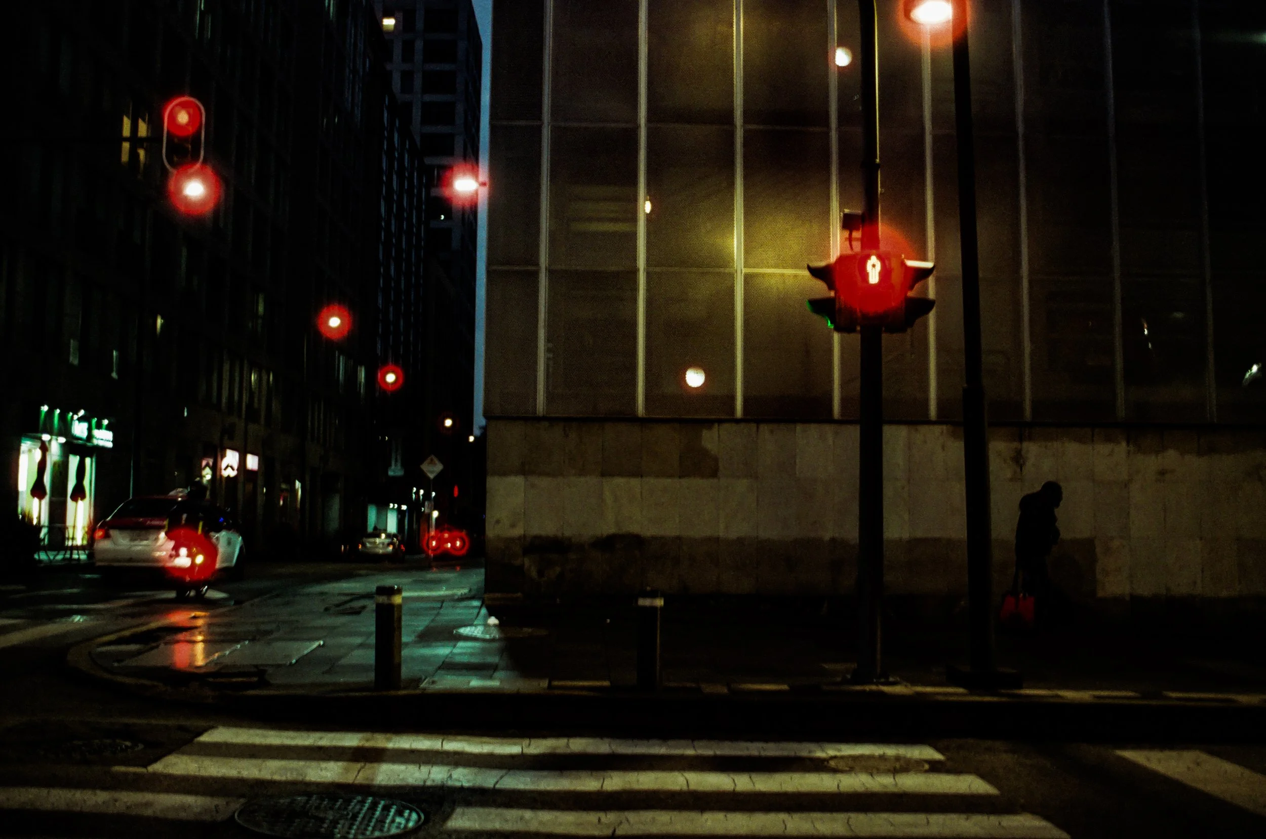 Nighttime street scene with red traffic lights, cars, and building facades, including a man walking on the sidewalk.