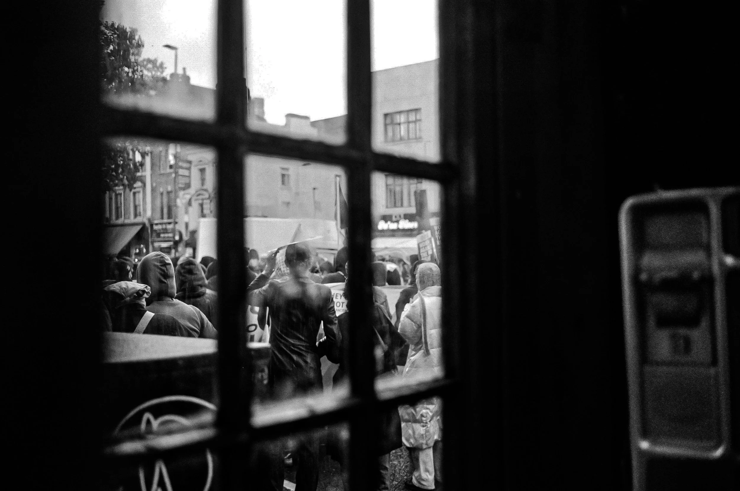 View of a crowd of people walking on a city street seen through window bars, with buildings and signs in the background.