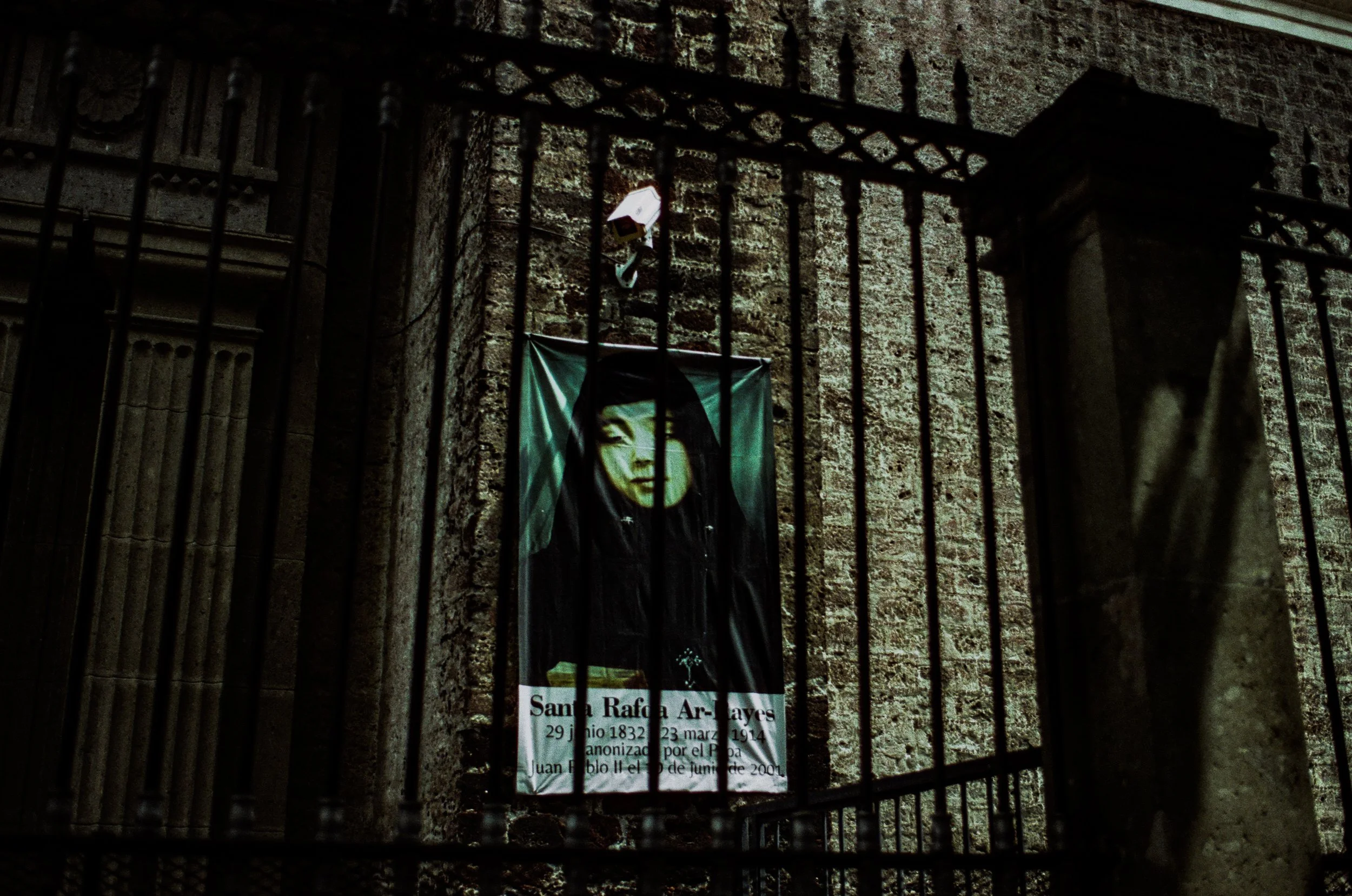 A barred window or gate with a religious poster of Santa Rafa Ar- Ayes, including dates, attached to a brick wall.