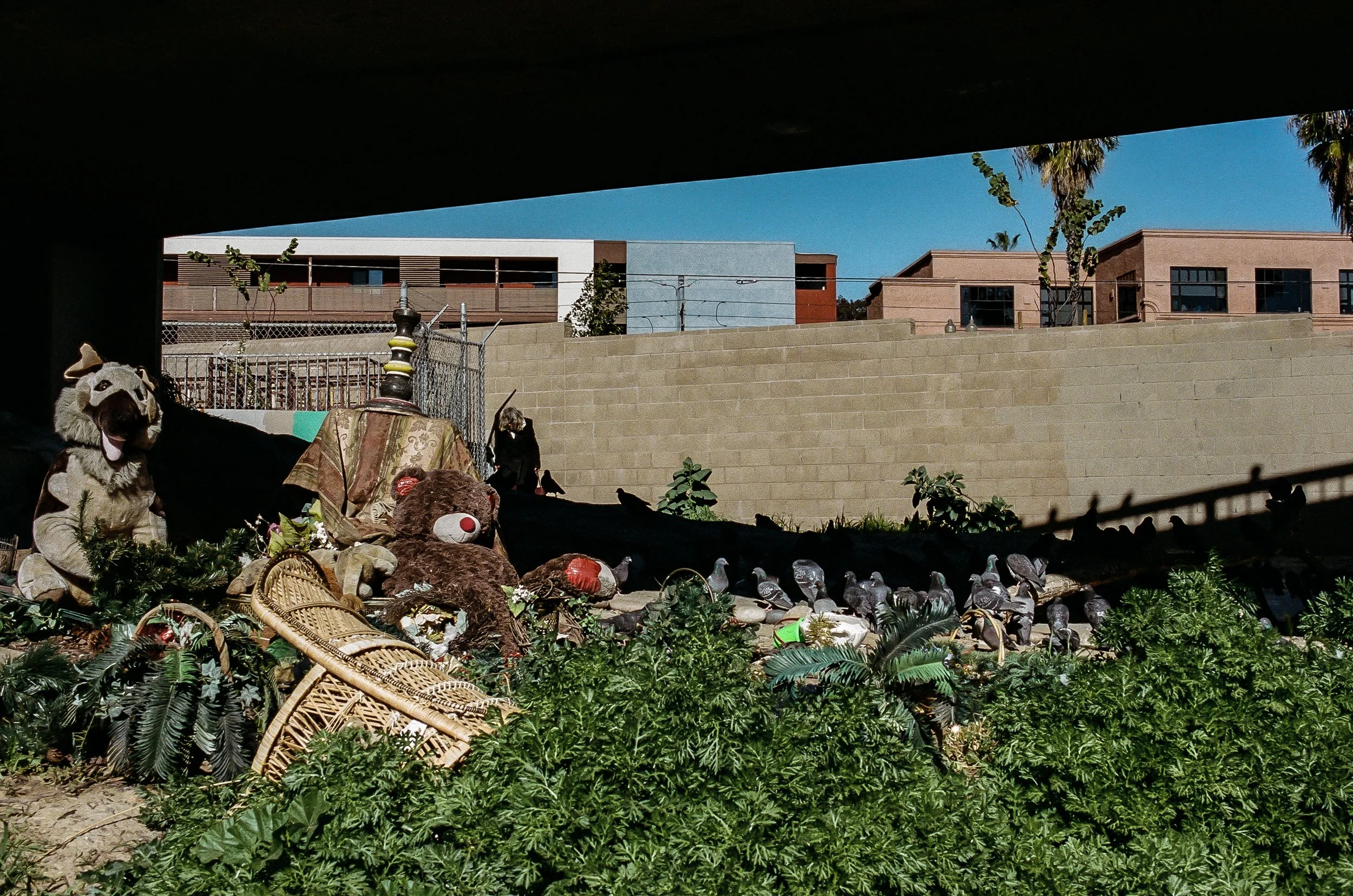 Various Halloween decorations including a teddy bear with a clown wig, a teddy bear with a red nose, and other stuffed animals, with greenery and wicker baskets in the foreground. In the background, there are residential buildings and a clear blue sk