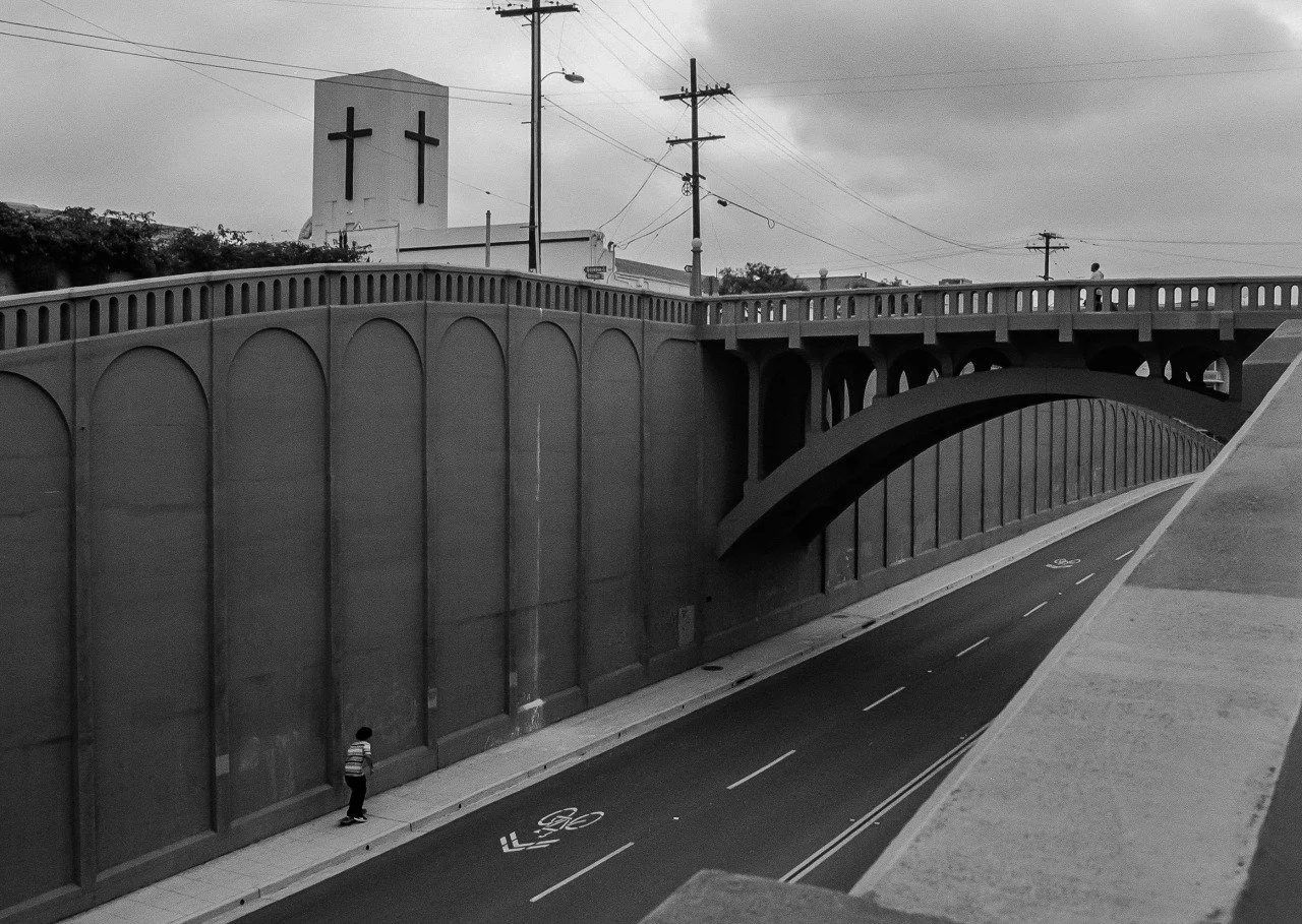 A black and white photo of a bridge with a person skateboarding underneath. The bridge has an arch and a wall with decorative arches. In the background, there is a church with two crosses on top and utility poles and wires.
