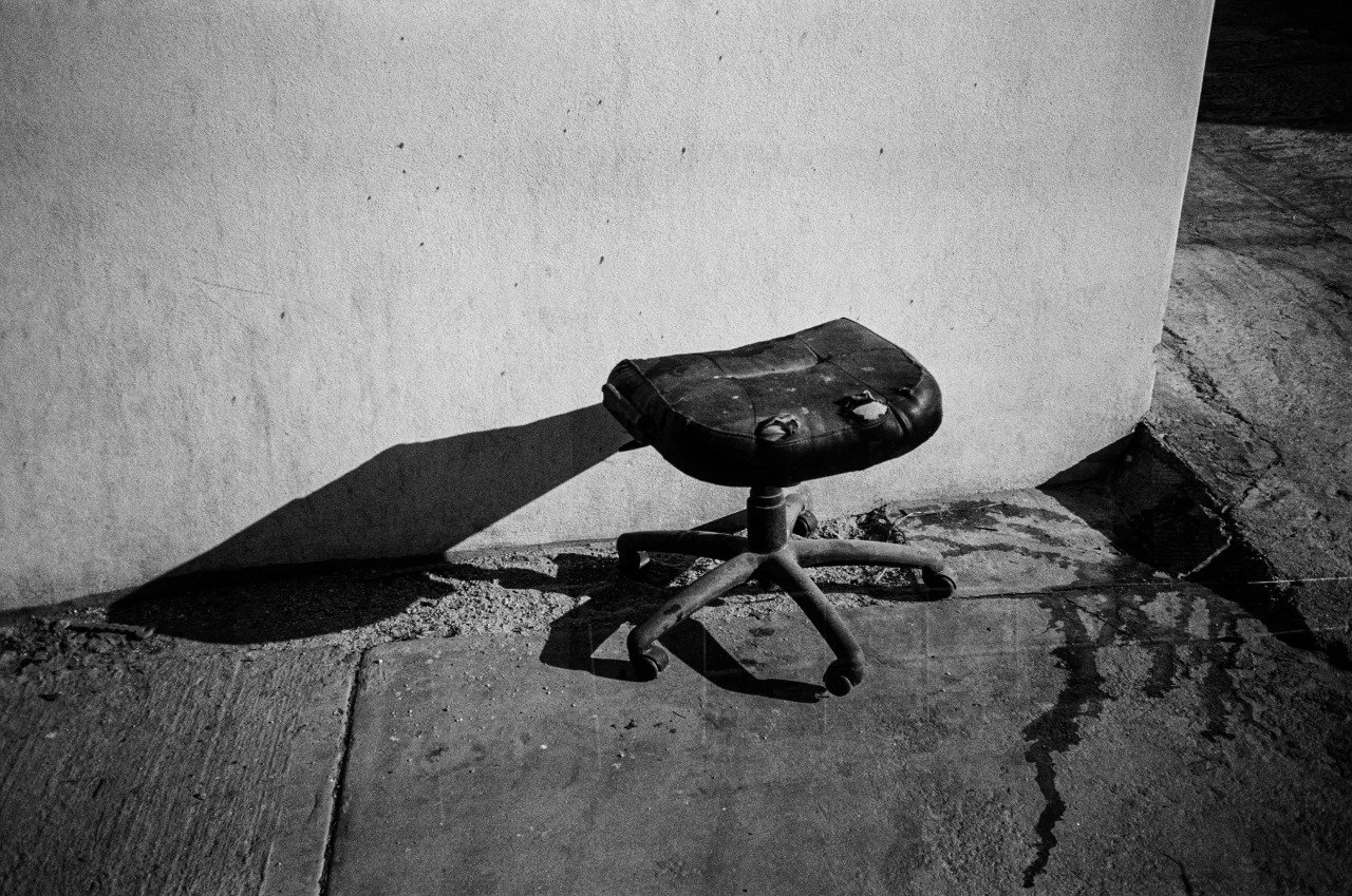 An old, worn office chair with a damaged black leather seat and a metal base, casting a shadow on a concrete sidewalk against a plain wall.