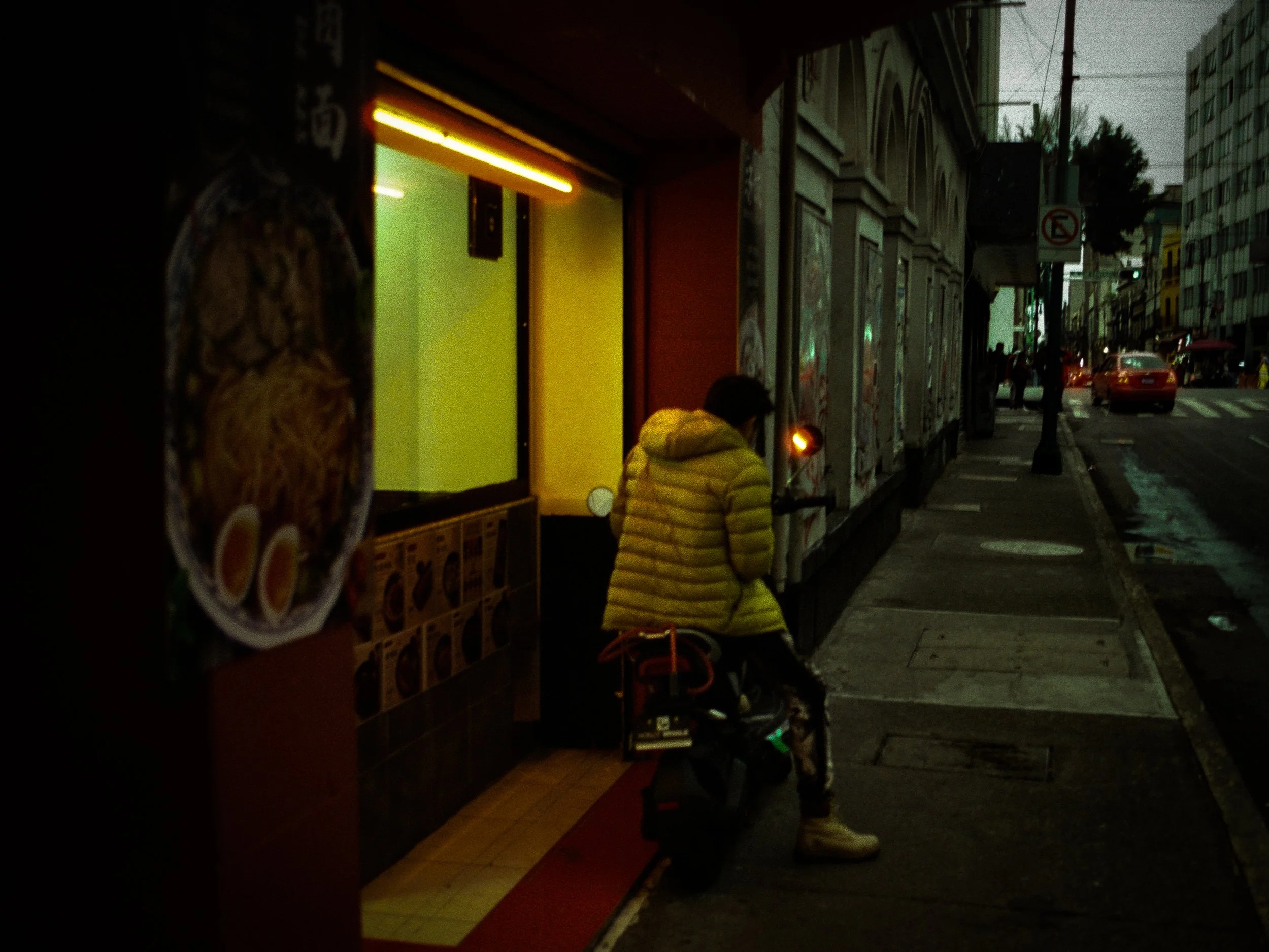 Nighttime street scene with a person in a yellow jacket looking at a phone outside a building, cars and pedestrians in the background, streetlights and traffic signals visible.