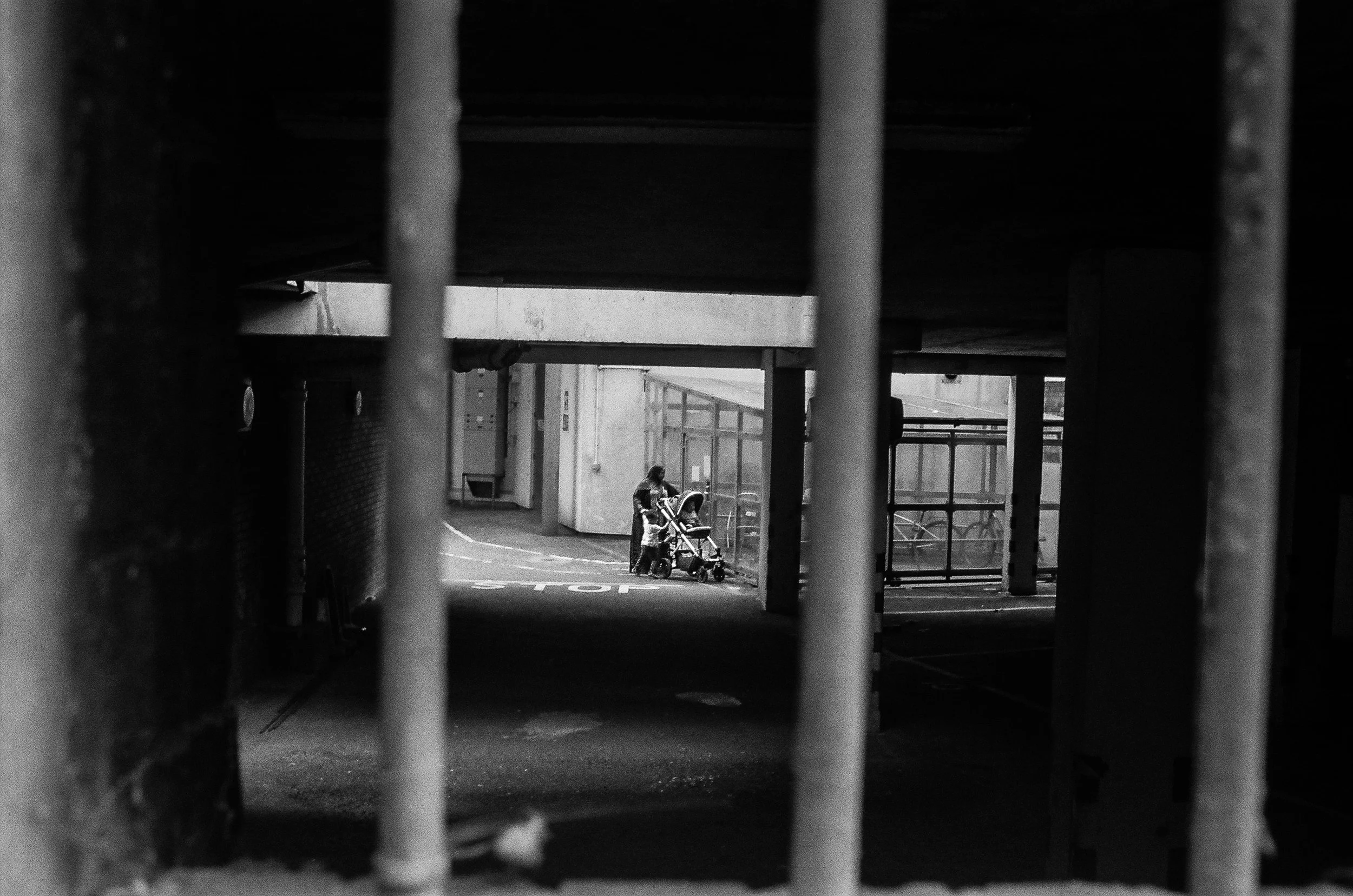A black and white photo taken through a metal fence showing a person with a stroller under an overpass or bridge, with urban buildings in the background.