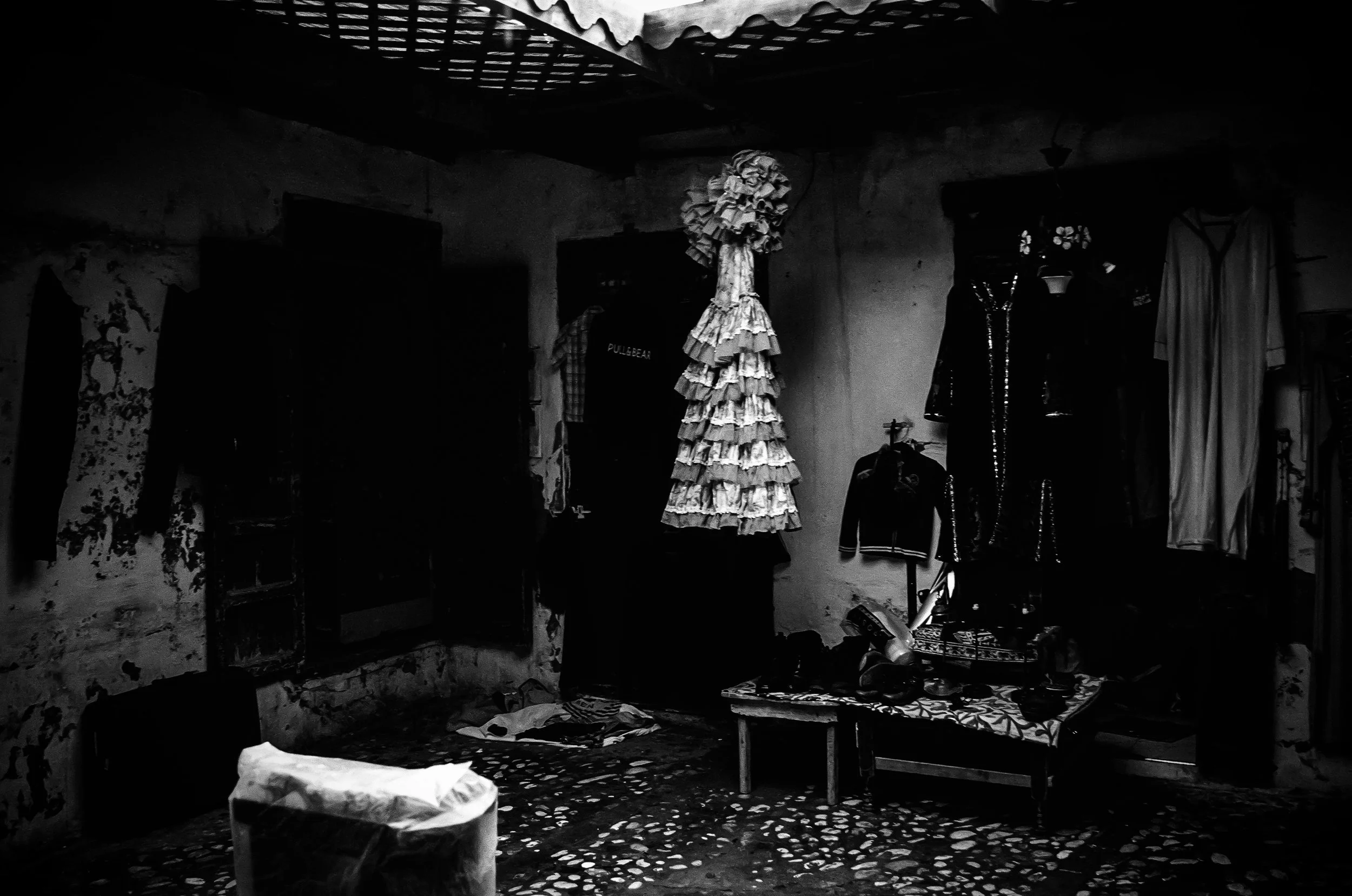 A black-and-white photo of a small, cluttered room with worn walls, a table, and various clothes hanging or stored. In the center, there is a dress form covered with layered fabric ruffles. The room appears to be a modest living or working space with