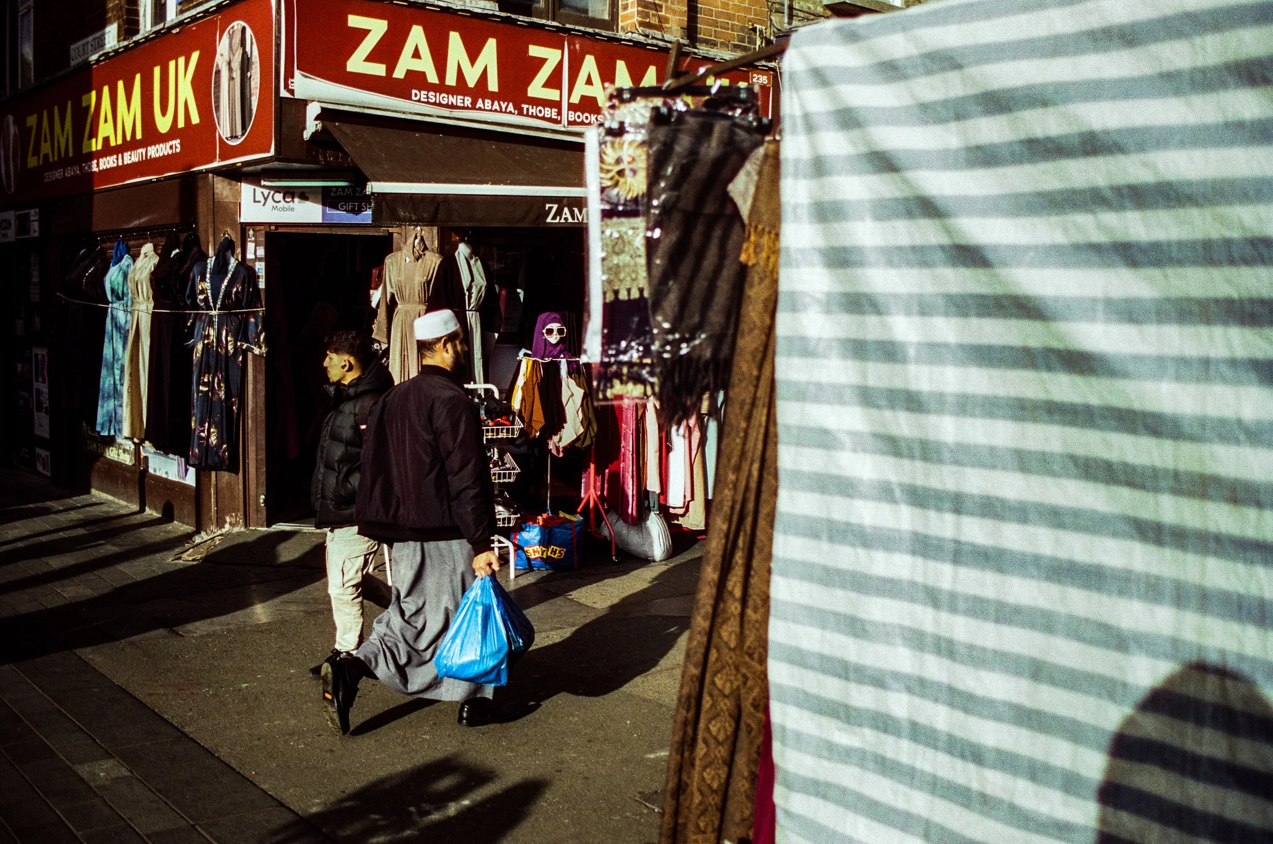Street scene outside a shop with a red sign reading 'ZAM ZAM UK' and displaying clothing on racks. Two men are walking past, one with a white cap, and the other wearing a black jacket and backpack, both carrying shopping bags.