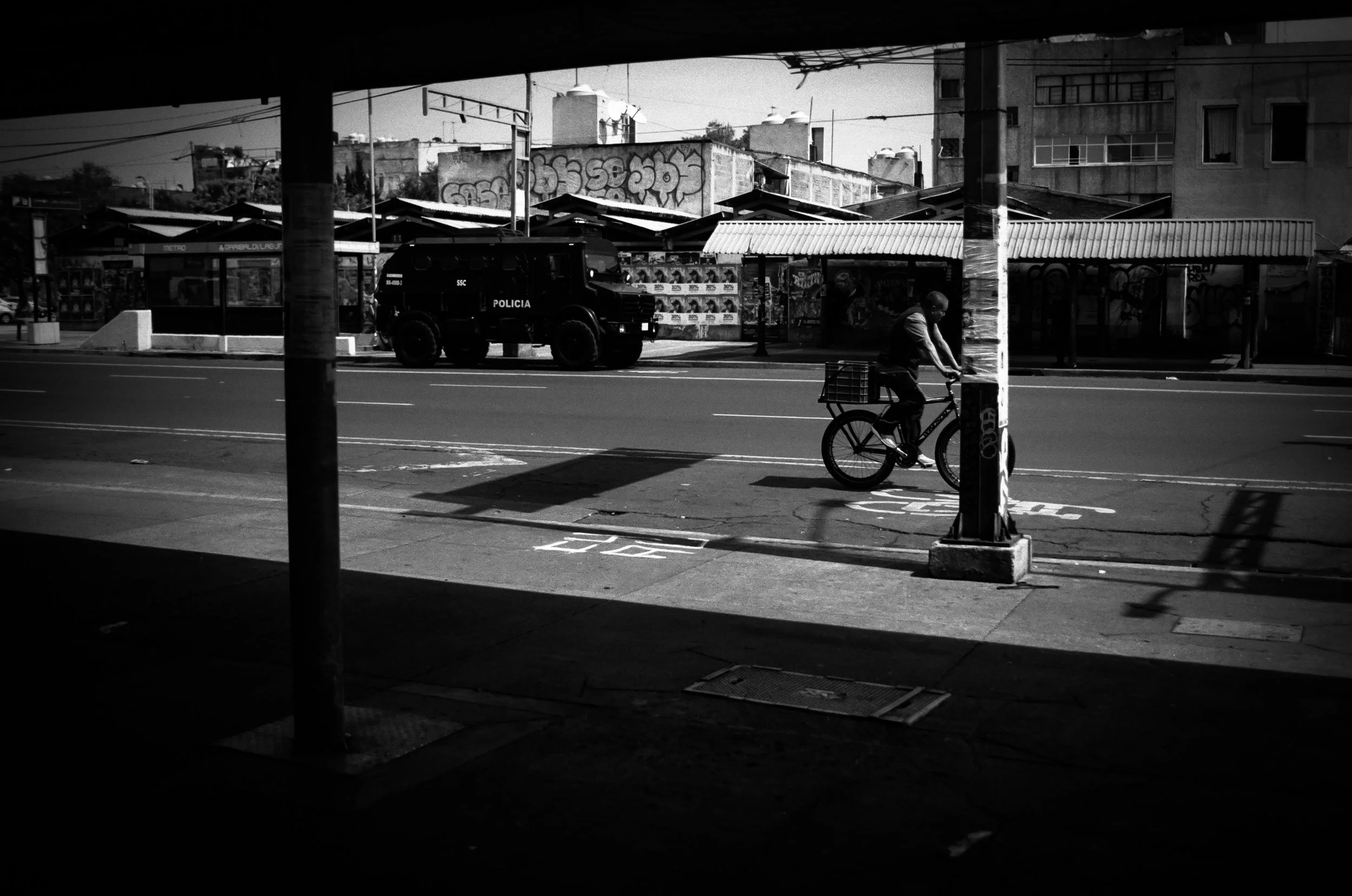 A black and white photo of an urban street scene viewed from inside a shaded area. A cyclist is riding along the street, and a police vehicle is parked across the road. There are multiple small shops with awnings and graffiti on nearby walls, with ap