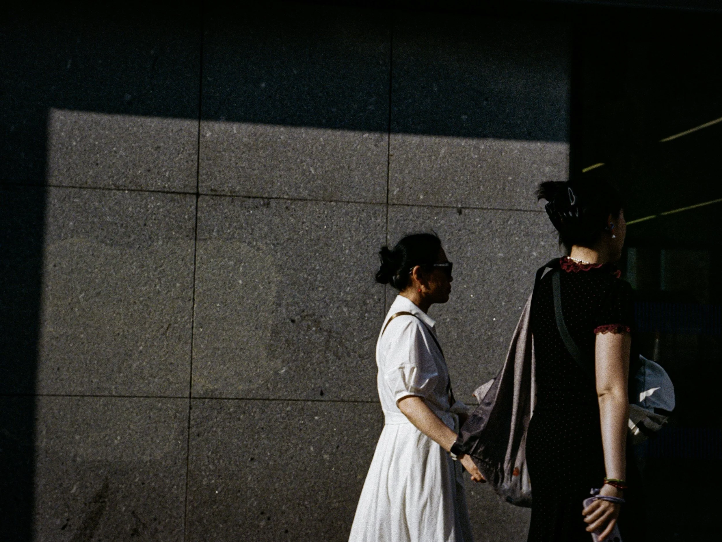 Side view of three women walking along a sidewalk, with a gray tiled wall behind them. The women are dressed in various outfits, some wearing dresses, with dark hair and accessories.