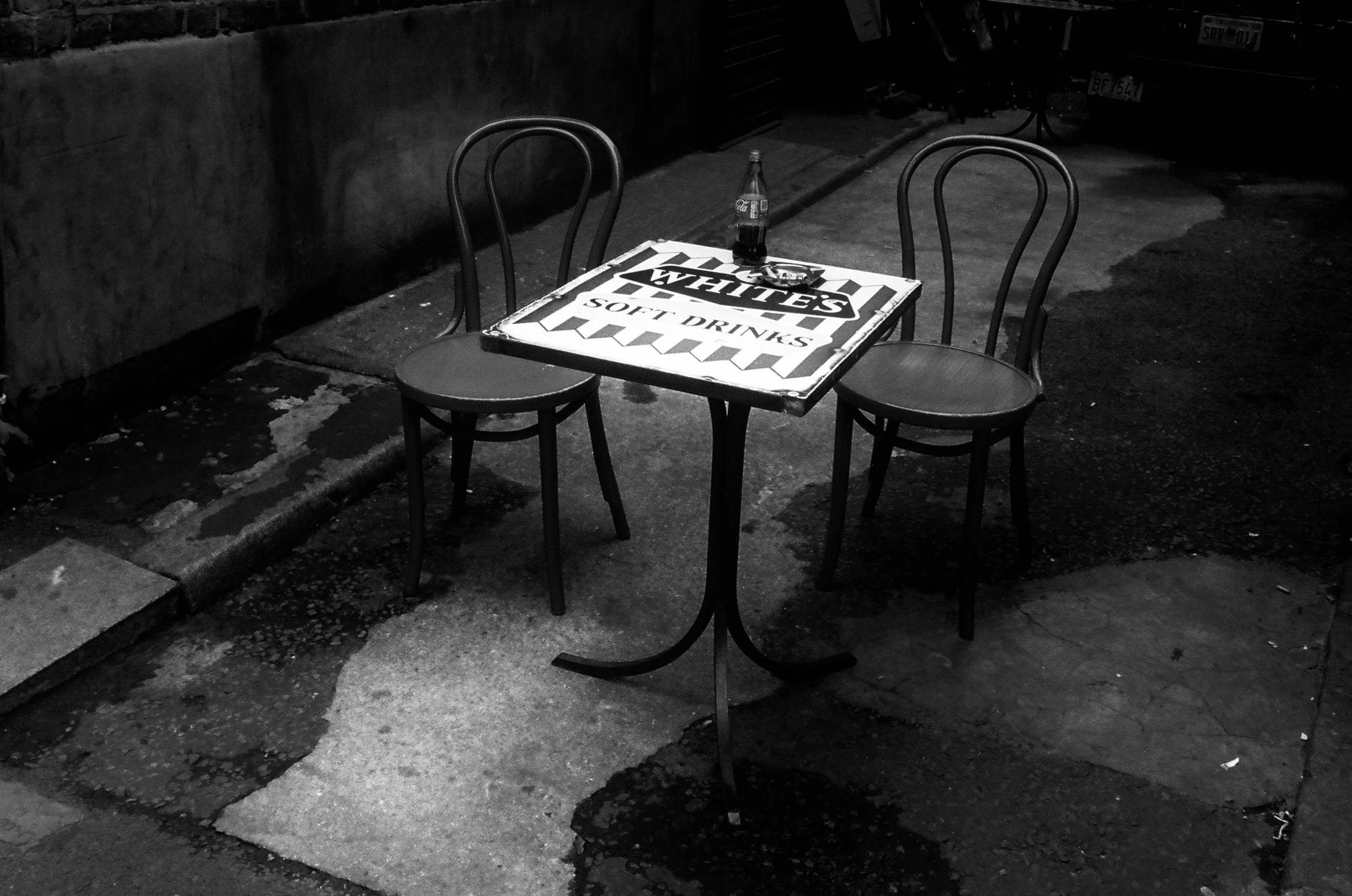 A small metal table with a sign that reads "WHITES SOFT DRINKS" under three chairs on a sidewalk at night, with a soda bottle placed on the table.