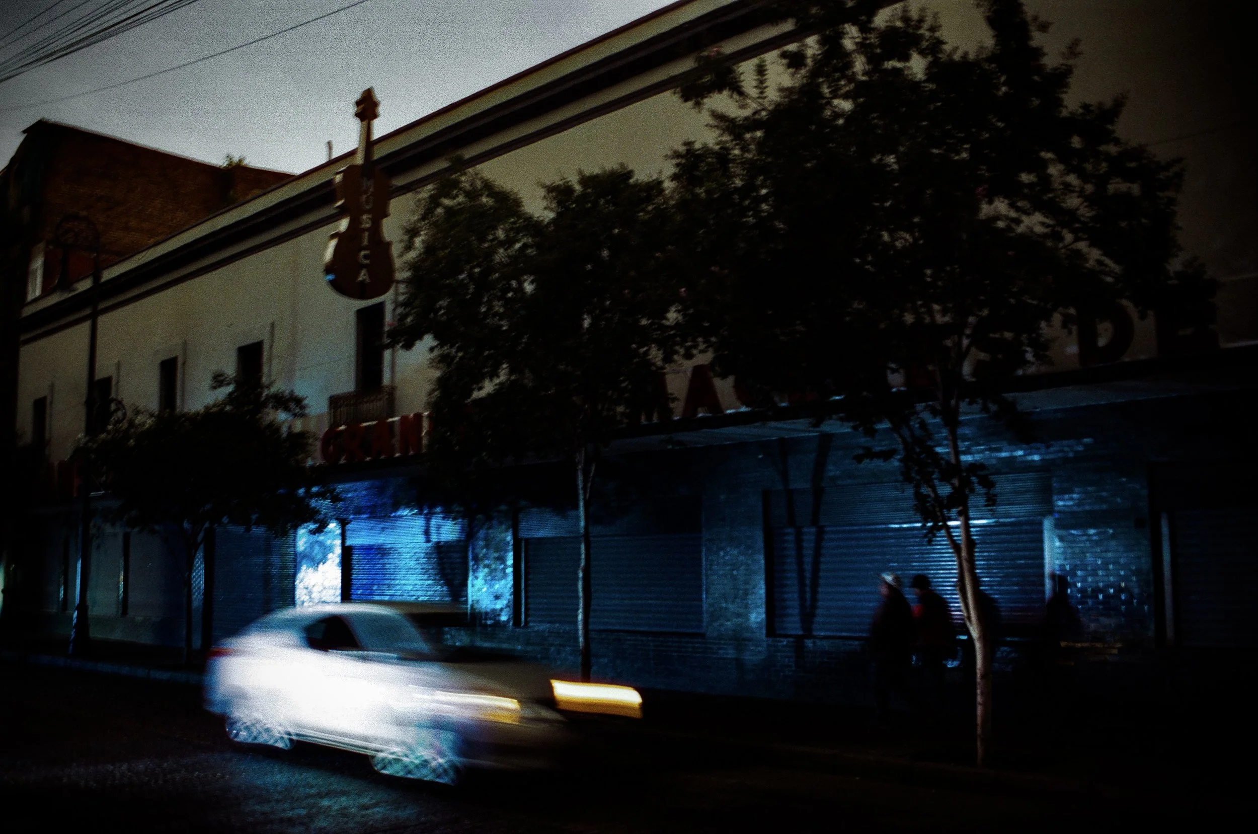 Night scene of a street with a building, trees, and a car passing by with motion blur. The building has a sign that reads 'TACIA' and appears to have a mural or painted advertisement on its wall. The sky is overcast or dark.