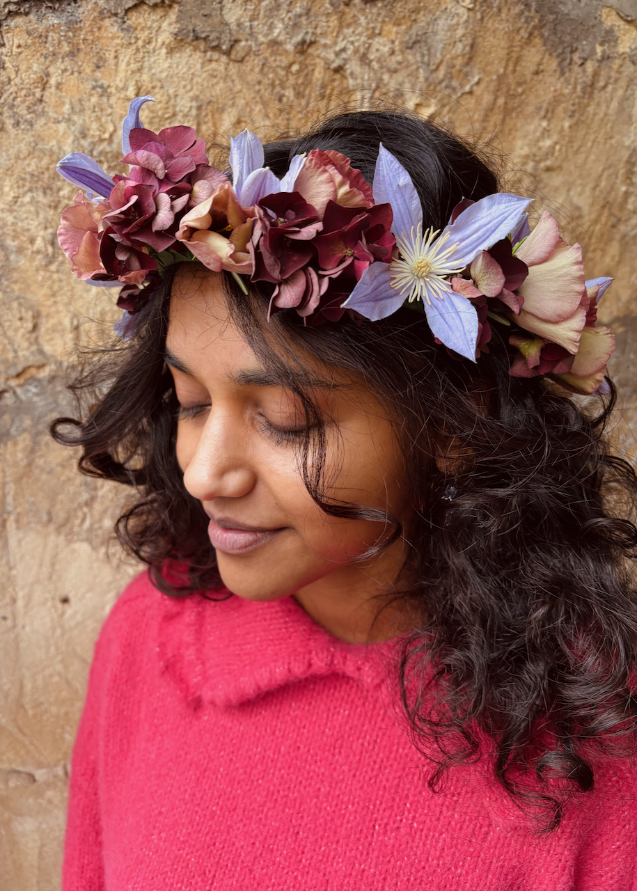 A woman wearing a floral crown with purple, pink, and cream flowers, dressed in a pink sweater with closed eyes and a peaceful expression, standing against a textured beige stone wall.