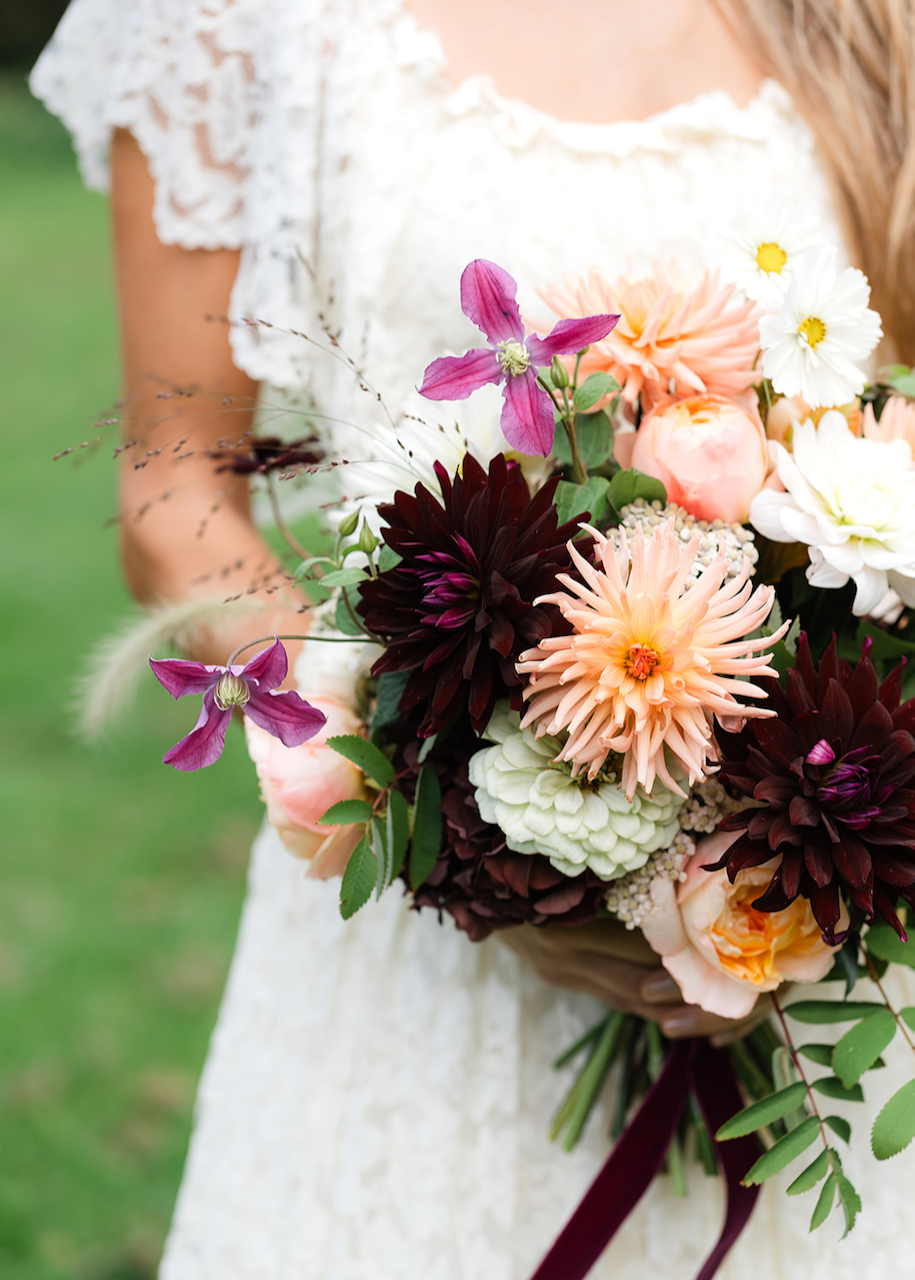 A woman in a white lace dress holding a bouquet of colorful flowers, including pink, purple, white, and dark red blooms.