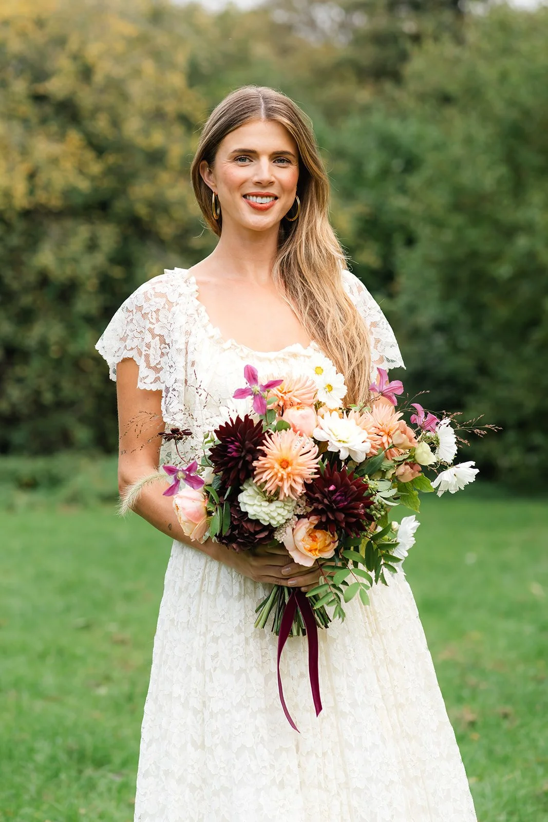 A woman in a white lace dress holding a colorful bouquet of flowers outdoors, smiling, with a background of green trees and grass.