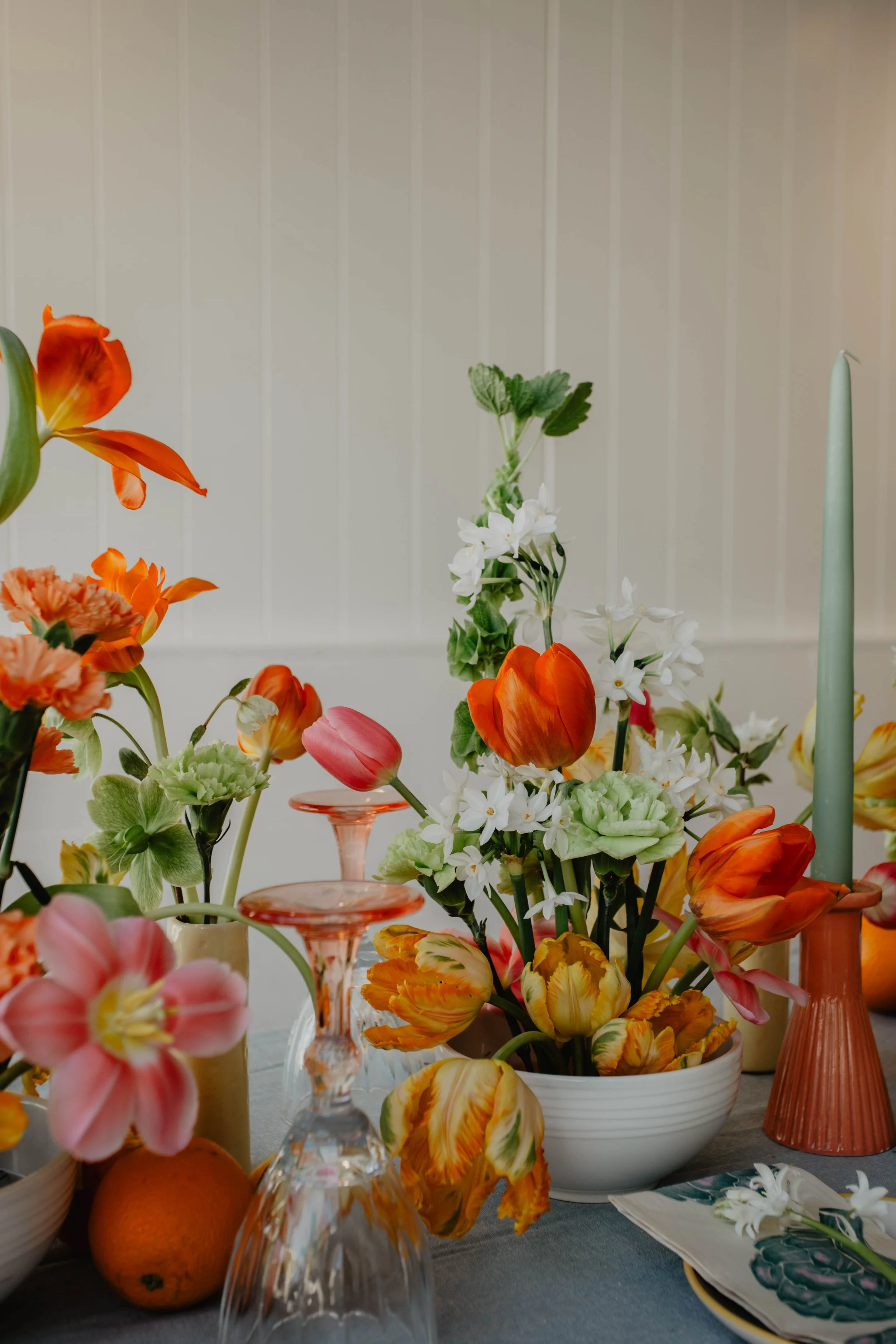 A floral arrangement with orange, pink, and white flowers in bowls and vases on a table, with candles and a gray tablecloth.