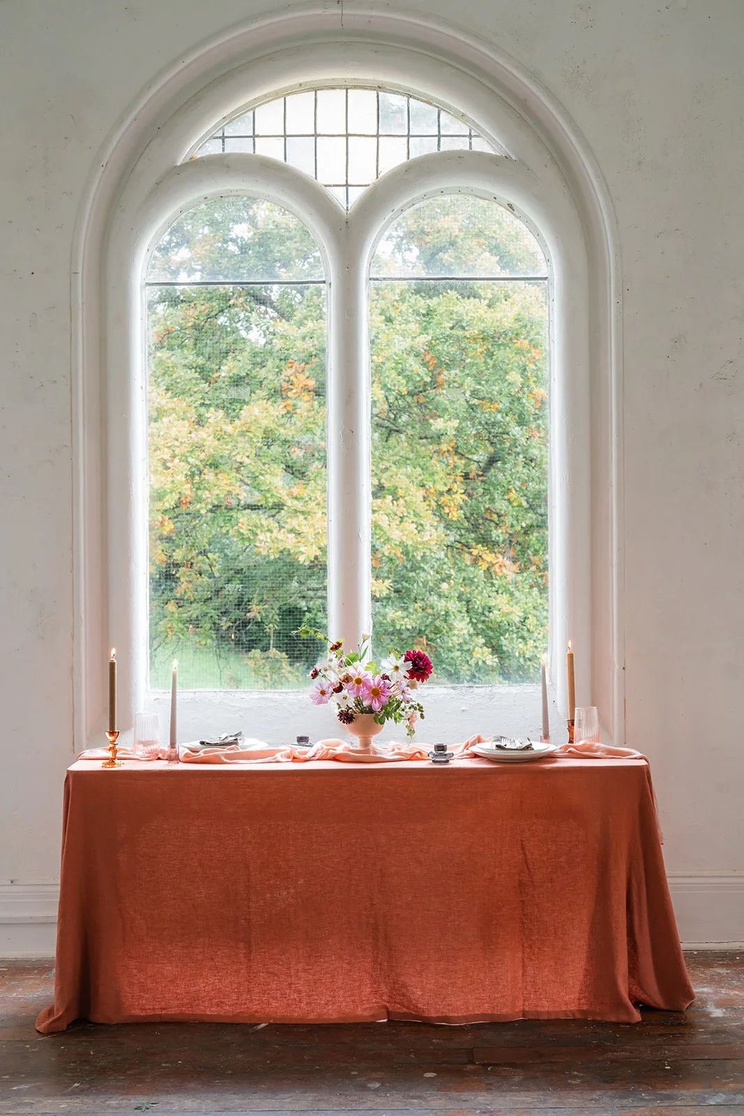A table with a peach-colored tablecloth set in front of a large window with three arched panes, decorated with a pink and white flower centerpiece, candlesticks, and place settings, overlooking autumn trees.