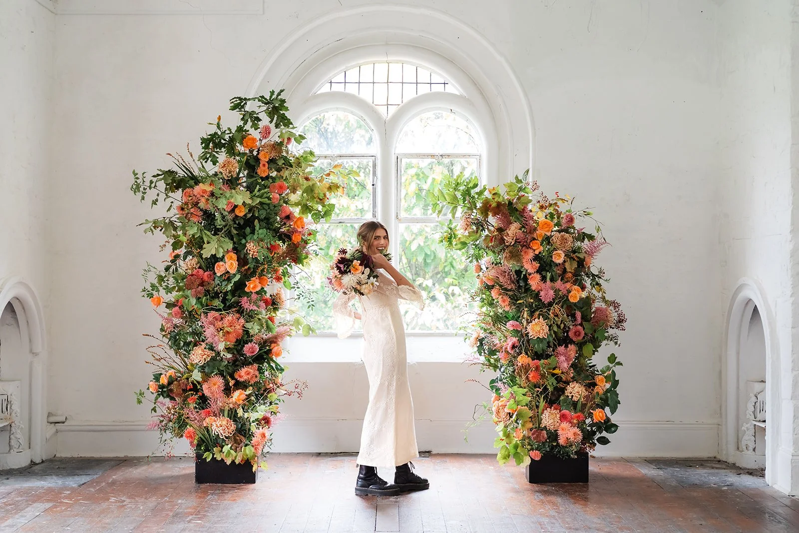 A woman in a white dress standing between two tall floral arrangements in black square pots, near a large arched window with greenery outside in the background.
