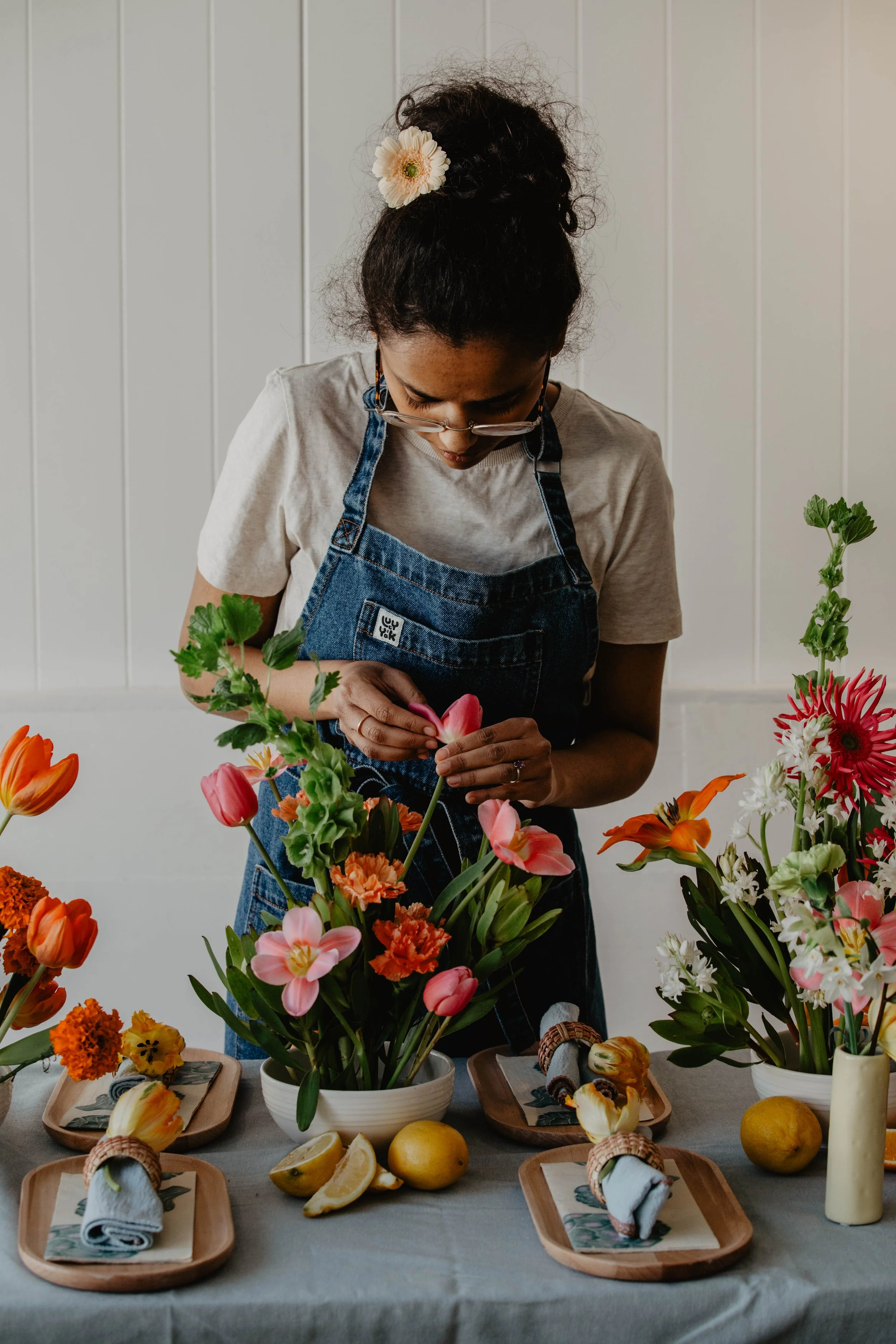 A woman arranging flowers on a table with various vases, lemons, and napkin settings.