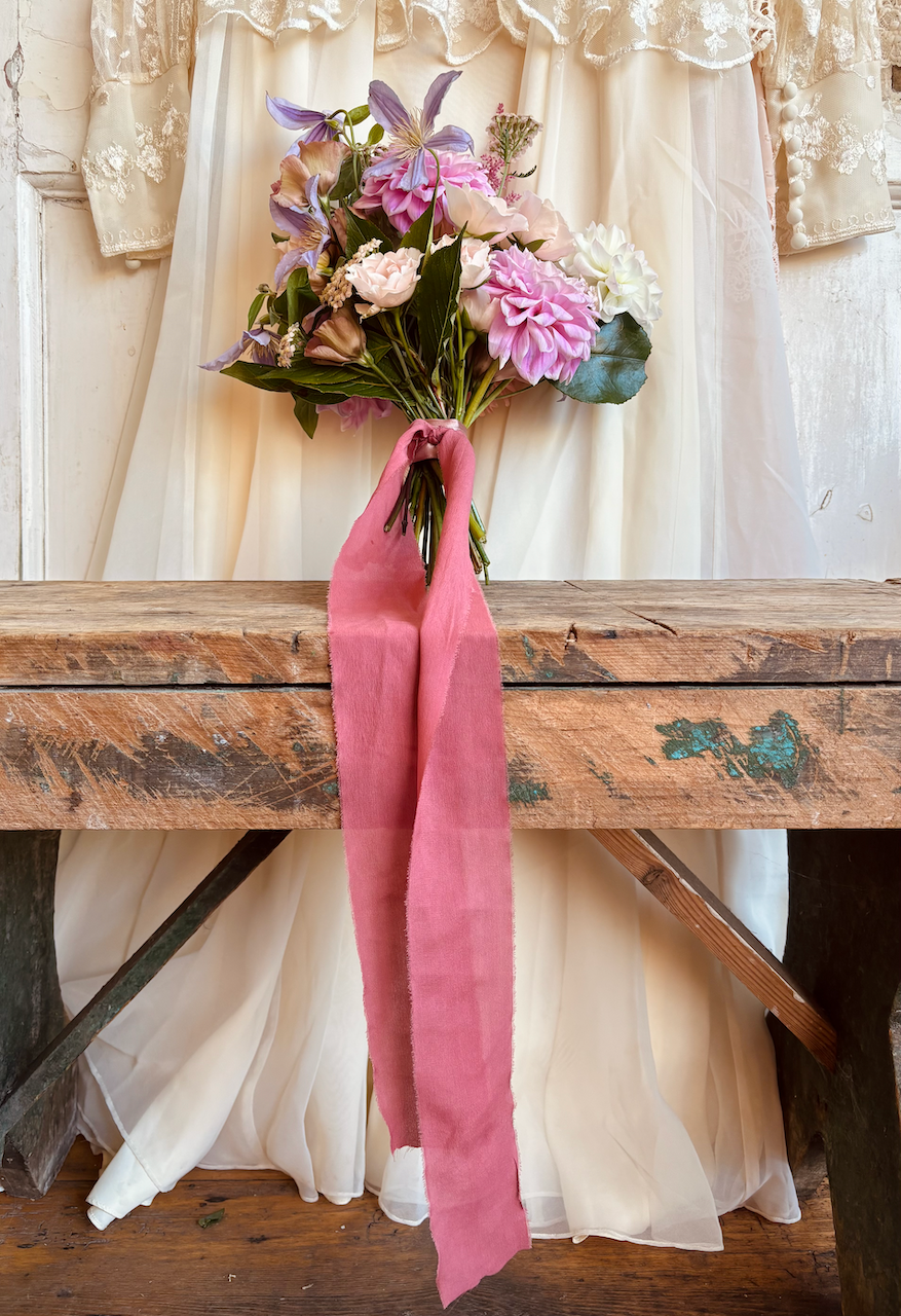 A colorful bouquet of pink, purple, white, and green flowers tied with a pink fabric ribbon rests on a rustic wooden table, with a vintage cream-colored curtain backdrop.