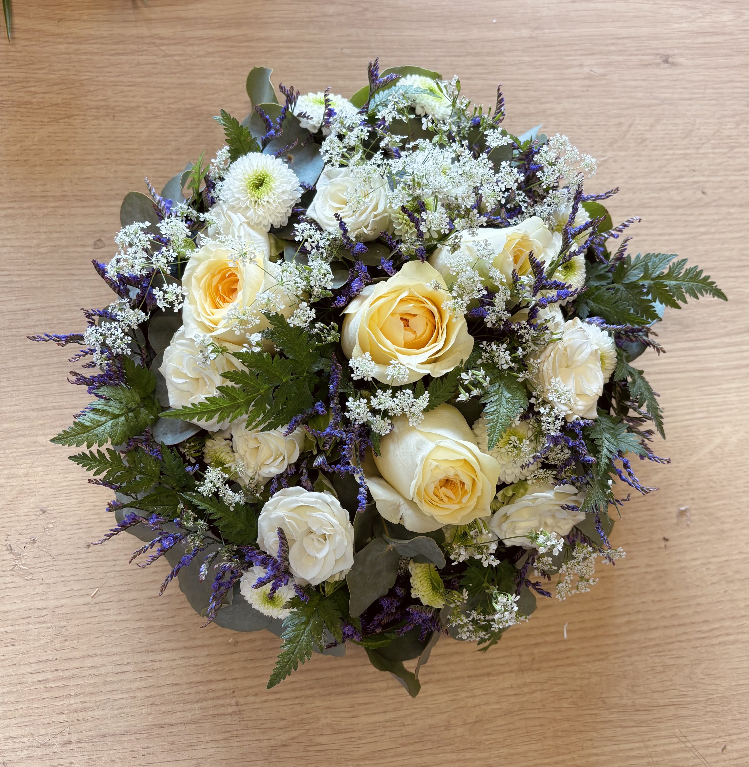 A round bouquet of white and cream roses, small white flowers, and purple accents, with green foliage, placed on a wooden surface.