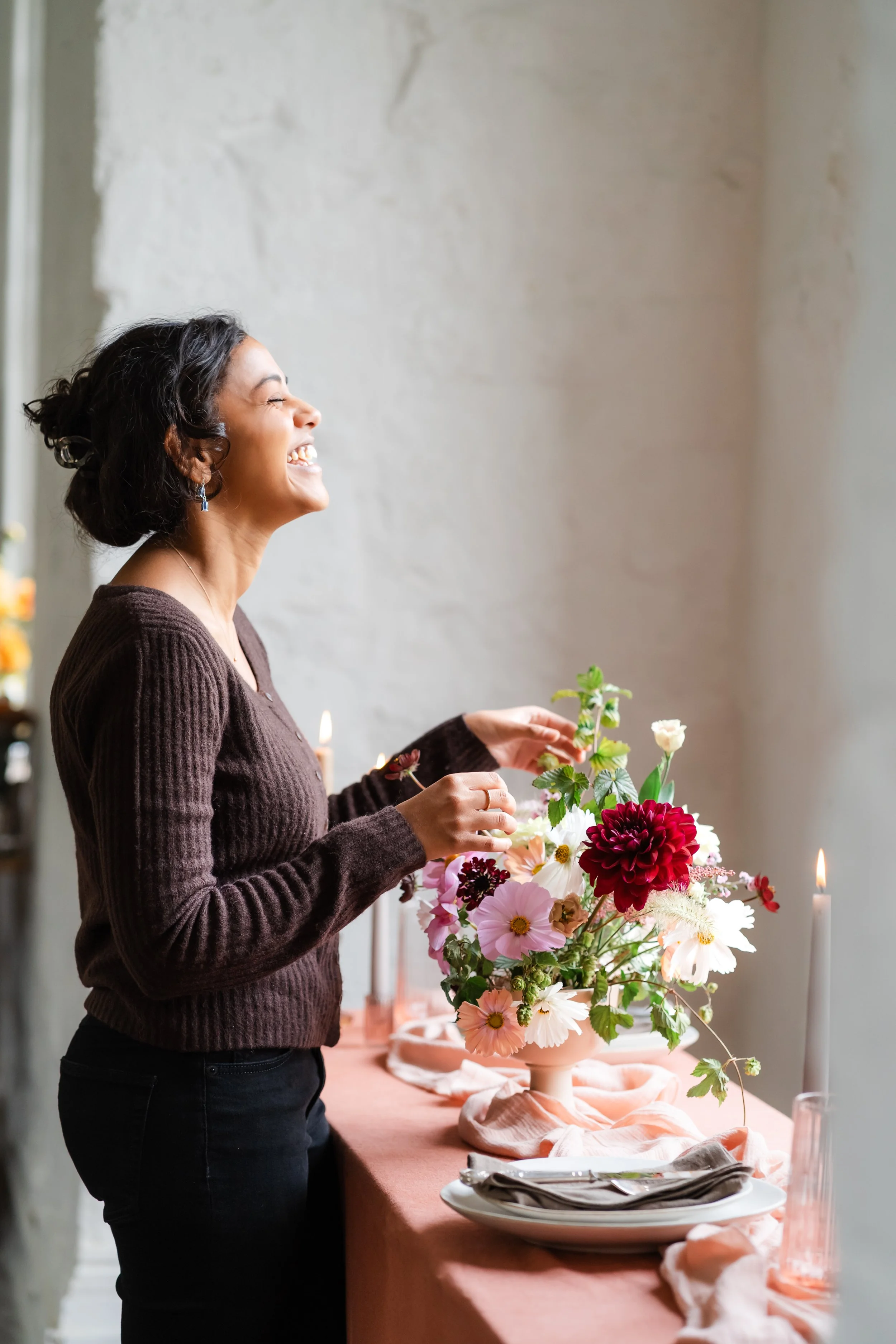 A woman with dark hair and earrings arranging a floral centerpiece on a table decorated with candles, a pink tablecloth, and place settings.