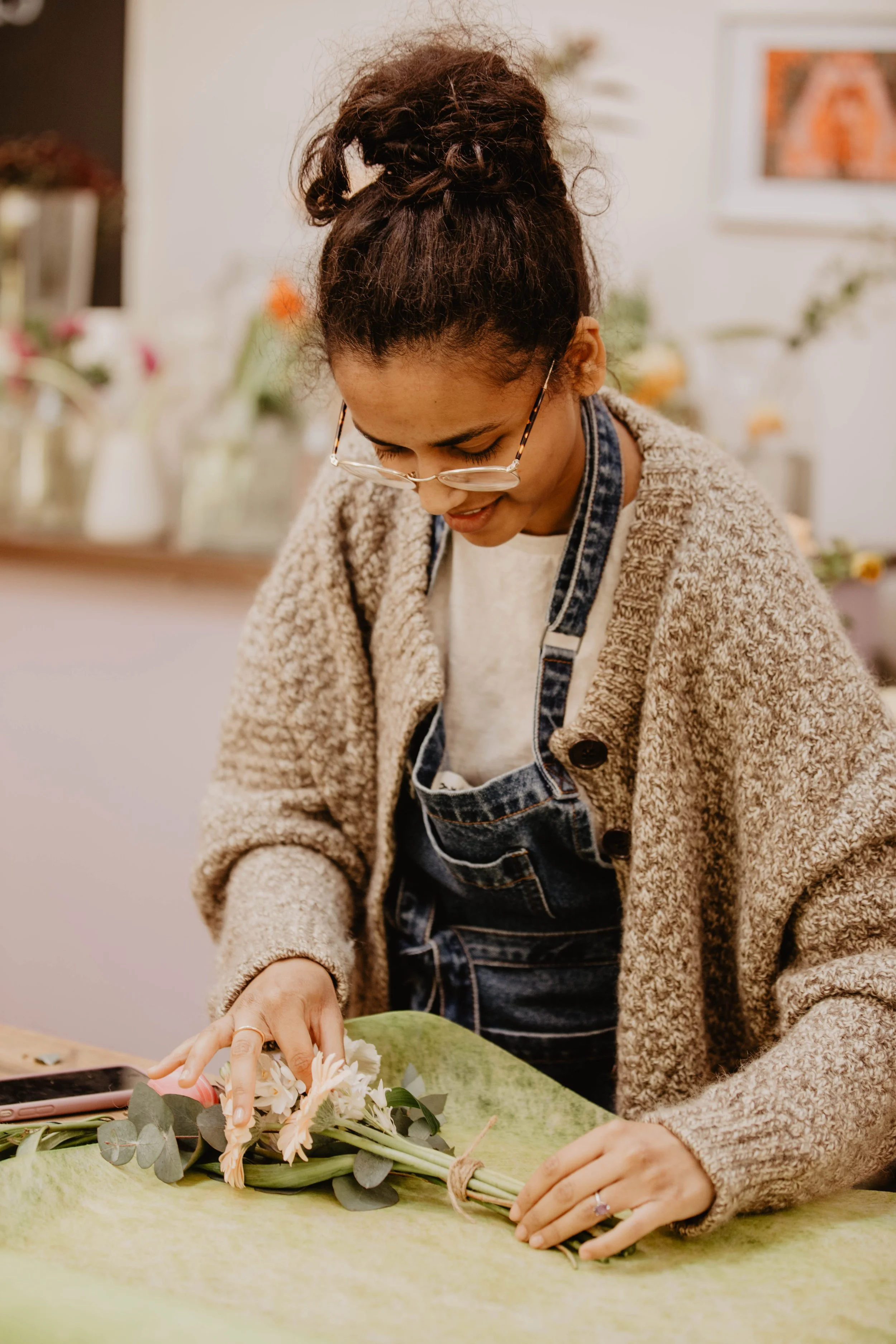 A woman with glasses and curly hair tied up in a bun is arranging a bouquet of flowers on a green surface. She is wearing a beige fuzzy sweater, white shirt, and denim overalls.