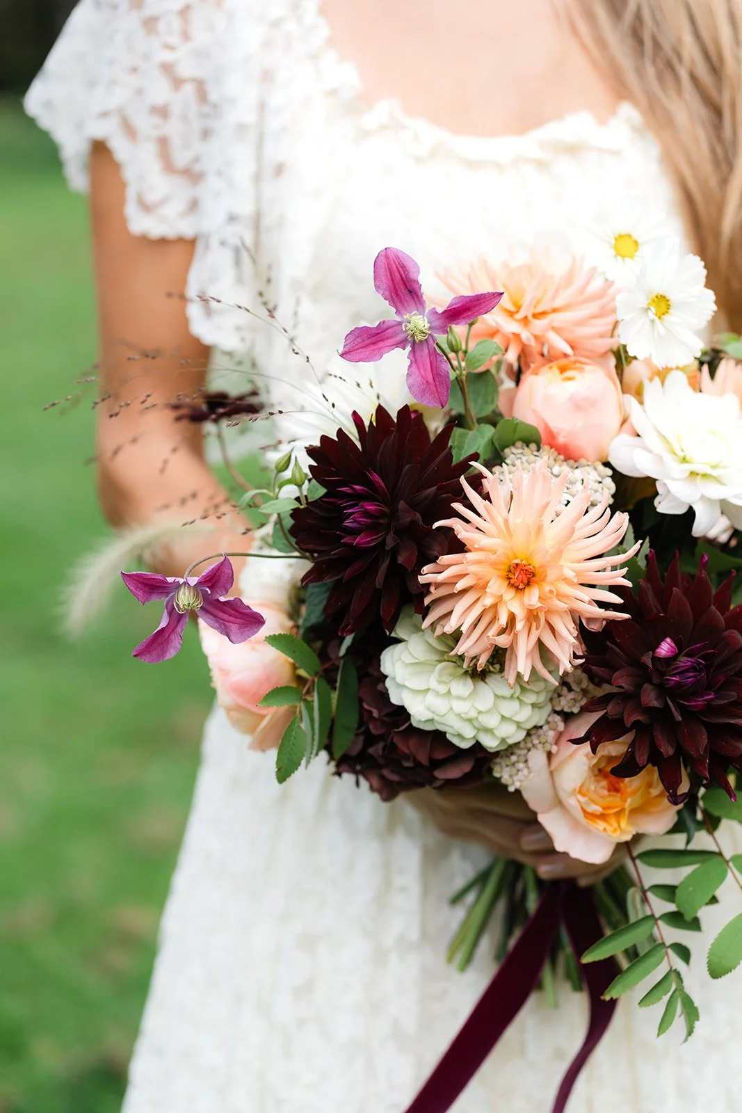 A woman in a white lace dress holding a colorful bouquet of flowers, including pink, white, and dark purple blooms.