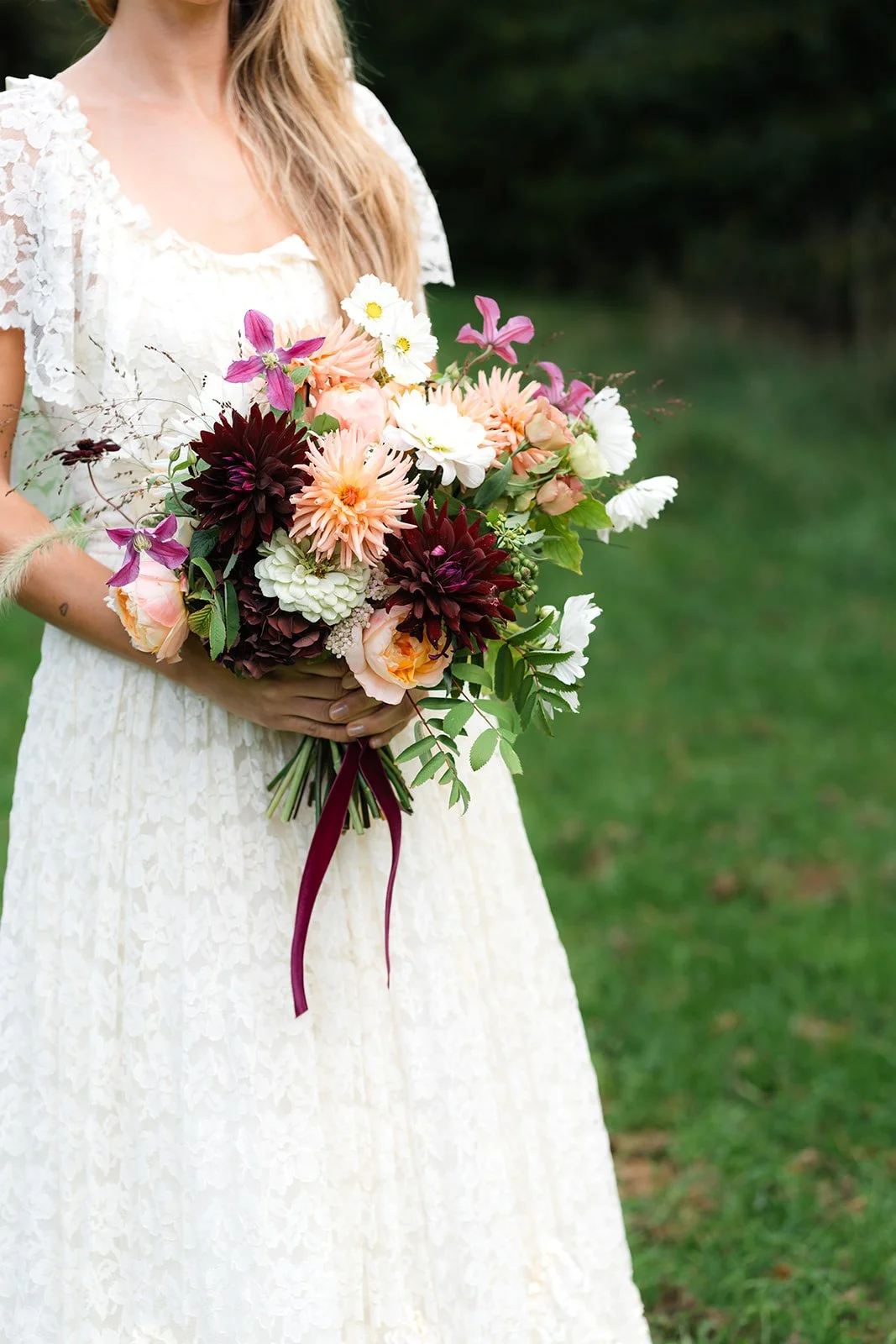 A woman in a white lace dress holding a large bouquet of assorted flowers, including pink, white, and dark red blooms, with a burgundy ribbon, standing on a grassy area.